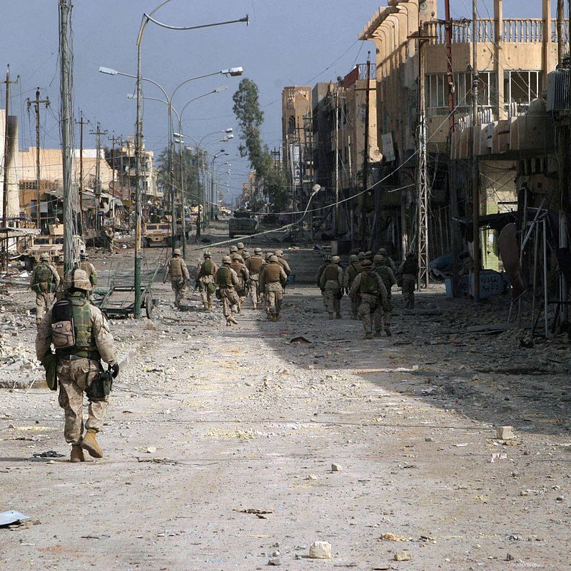 تظهر الصورة مشهدًا لمدينة مدمرة، حيث تسير مجموعة من الجنود في شارعٍ واسع. الشوارع تبدو خالية وتظهر فيها آثار الدمار، مثل المباني المتضررة والأدوات المتروكة. السماء تبدو غائمة، مما يضفي على المشهد طابعًا قاتمًا. الجنود يرتدون زيًا عسكريًا ويحملون معداتهم، مما يشير إلى أنهم في مهمة عسكرية.