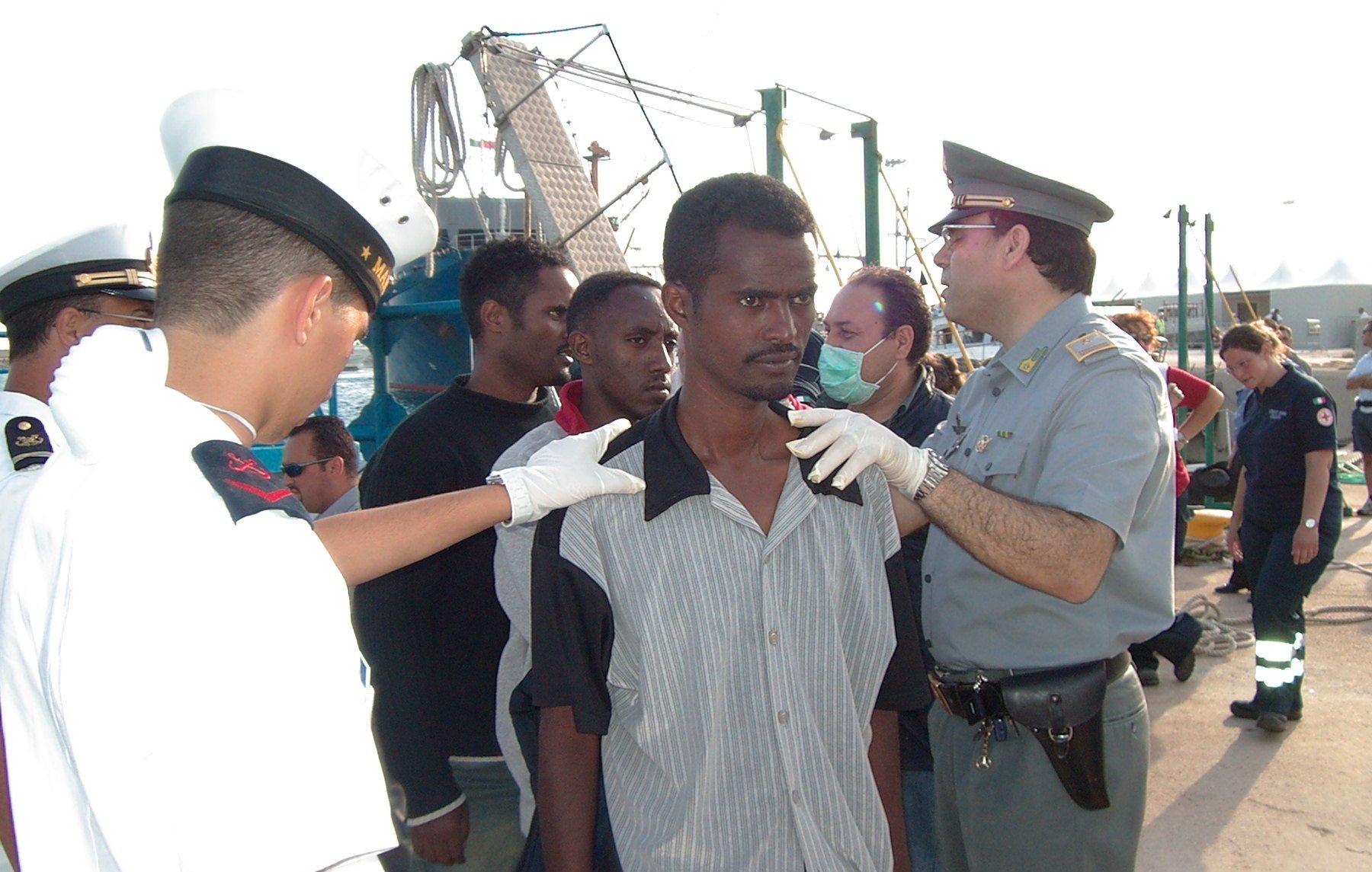 L'image montre une scène avec plusieurs personnes. Au premier plan, un homme se tient debout, apparemment en train d'être examiné par des agents portant des uniformes. Il y a un groupe d'autres personnes en arrière-plan, semblant également être supervisées. L'environnement suggère que cela se passe dans un port ou une zone maritime, avec des bateaux et des structures portuaires visibles. Les agents semblent s'occuper de la situation de manière attentive.