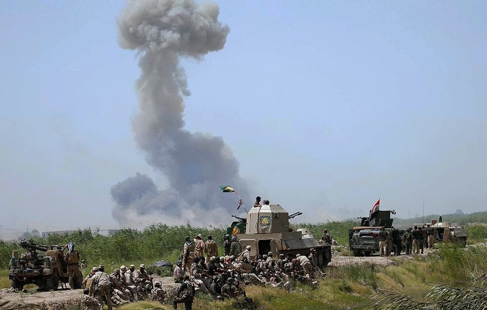 L'image montre un paysage militaire avec des soldats au sol et des véhicules blindés. En arrière-plan, une épaisse colonne de fumée s'élève dans le ciel, suggérant une explosion ou un feu. Les soldats semblent se regrouper, certains assis au sol, tandis que d'autres sont postés près des véhicules. L'environnement est rural, avec une végétation légèrement visible. L'atmosphère semble tendue en raison de la situation militaire représentée.