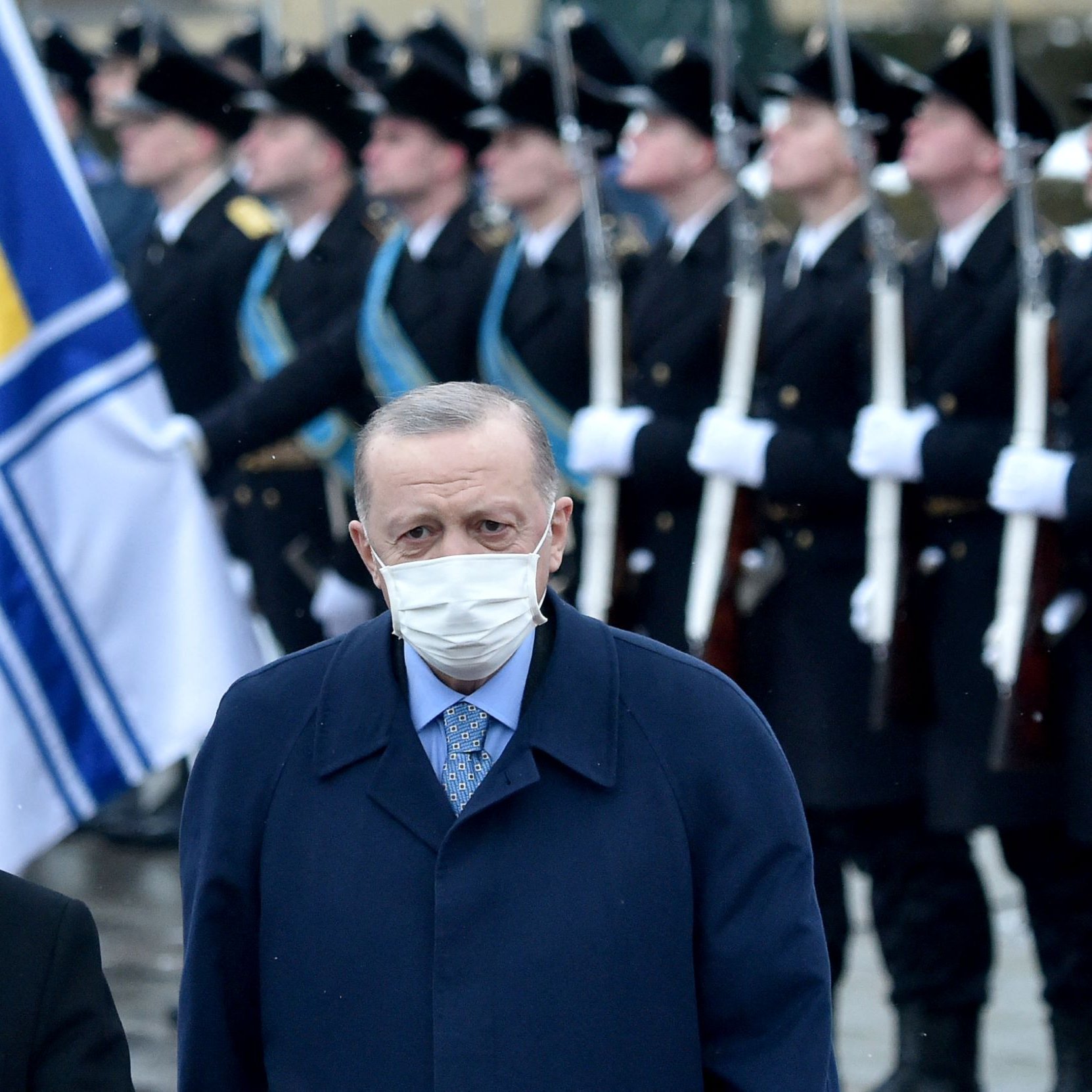 در تصویر، دو مرد در پیش‌زمینه دیده می‌شوند که هر دو ماسک زده‌اند. یکی از آن‌ها ظاهراً مقام رسمی است و دیگری نیز به نظر می‌رسد که رهبر یک کشور باشد. در پس‌زمینه، یک رژه نظامی در حال برگزاری است و سربازان با یونیفورم‌های رسمی و پرچم‌هایی در دست ایستاده‌اند. فضای کلی تصویر رسمی و جدی به نظر می‌رسد.