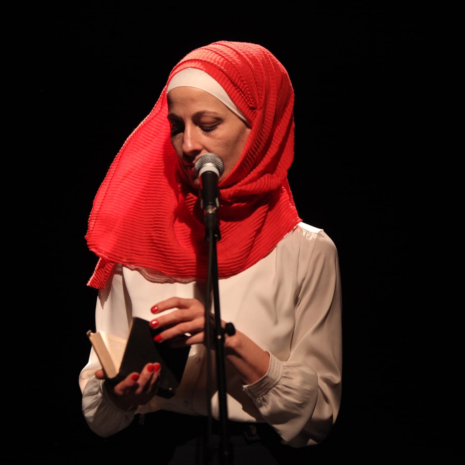 L'image montre une femme debout devant un microphone sur scène. Elle porte un hijab rouge vif et une blouse blanche. Elle tient un petit livre dans ses mains, semblant s'apprêter à lire ou à partager un texte. L'éclairage est sombre, mettant en valeur son visage et l'objet qu'elle tient. L'atmosphère semble intime et concentrée.