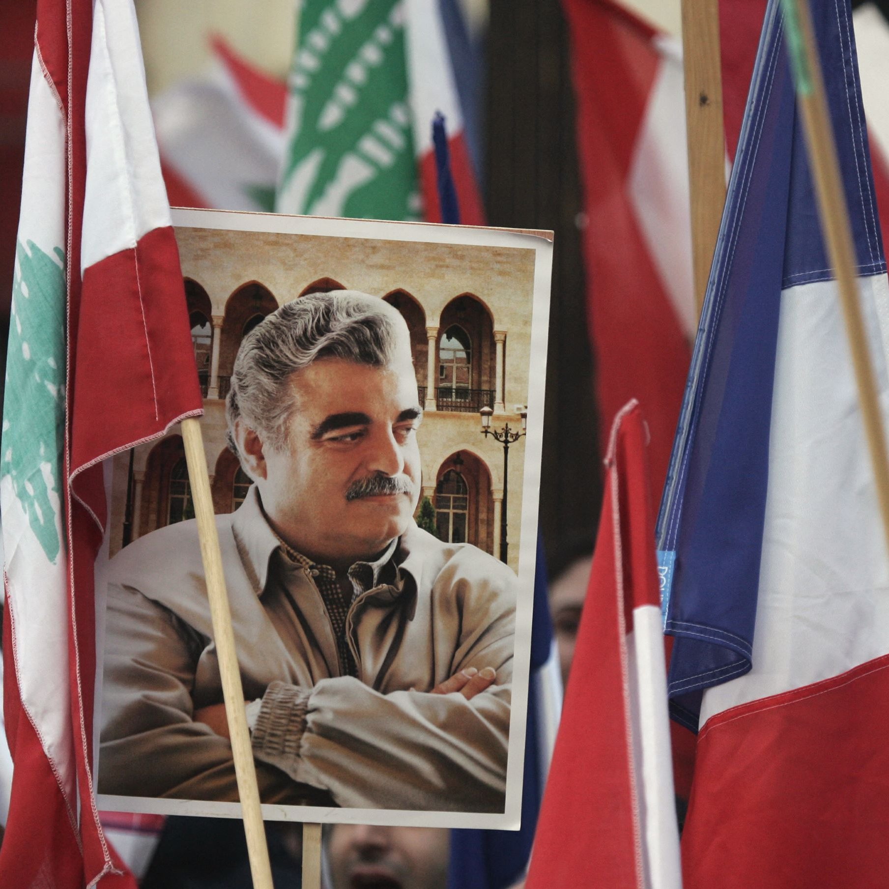L'image montre une manifestation où plusieurs drapeaux, dont ceux du Liban et de la France, sont visibles. Au centre, il y a un portrait d'un homme, tenant une expression sérieuse, et entouré de personnes tenant des drapeaux. L'atmosphère semble chargée d'émotion et d'engagement.