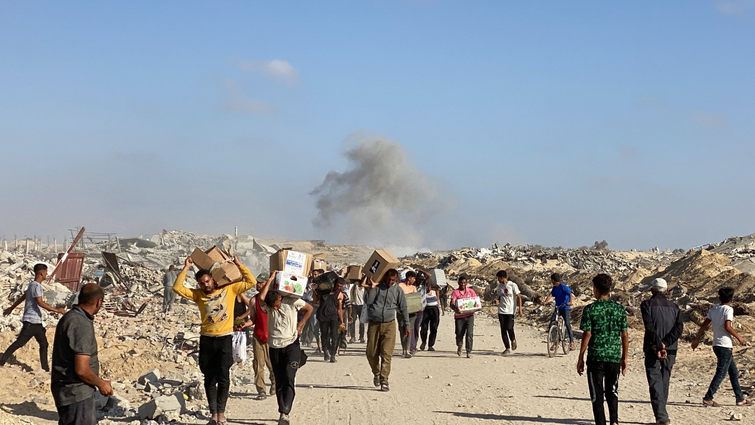 Des personnes marchent dans des décombres, portant des colis, avec de la fumée au loin.