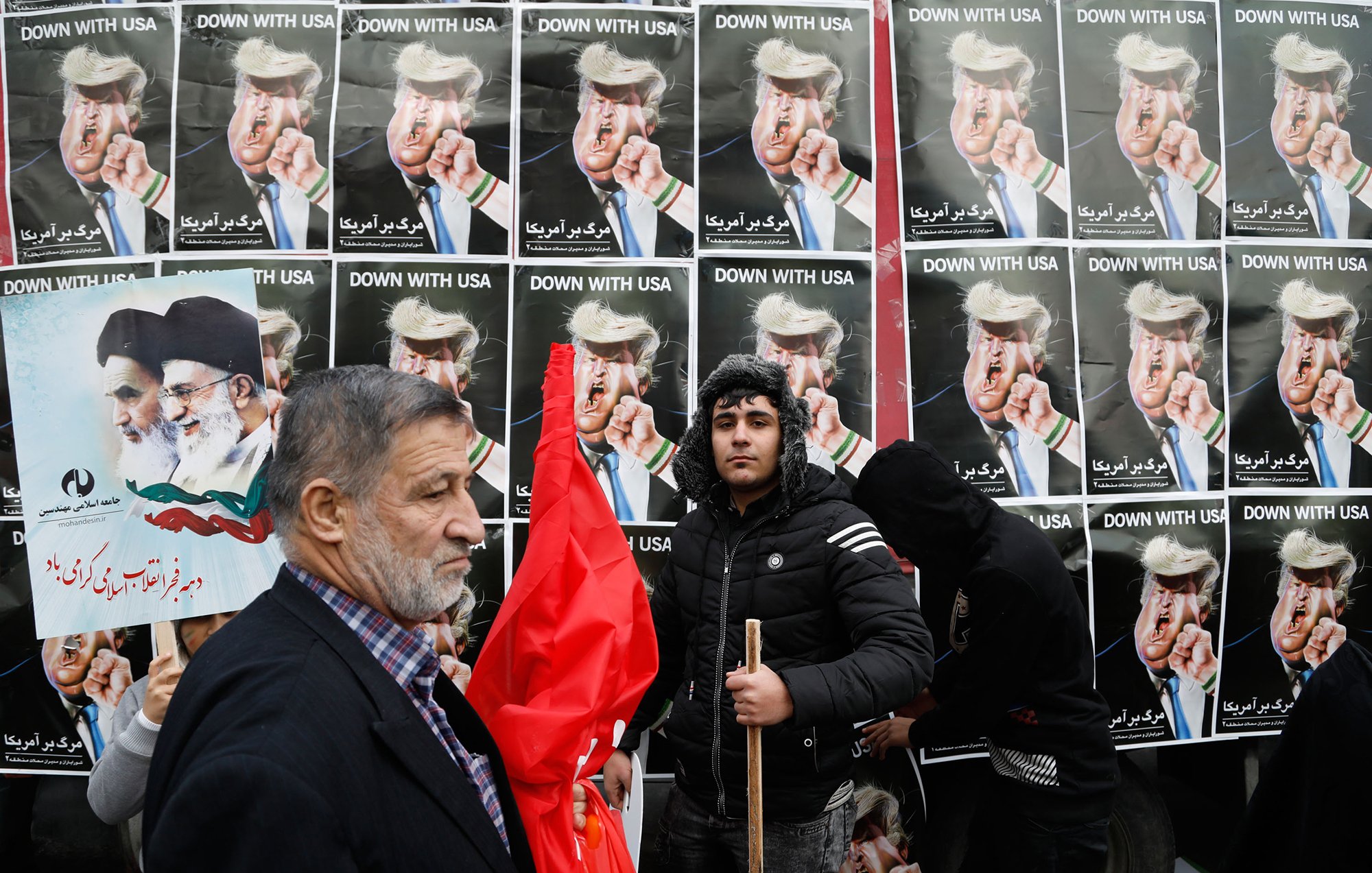 در این تصویر، گروهی از مردم در حال تظاهرات به نمایش گذاشته‌اند. در پس‌زمینه، پوسترهایی با تصاویر رهبران سیاسی و متنی به زبان فارسی دیده می‌شود که به موضوعی خاص اشاره دارد. یکی از افراد به سمت دوربین نگاه می‌کند و فرد دیگری با یک چوب در دست و لباسی سیاه در کنار او ایستاده است. برخی از افراد دیگر نیز در این صحنه حضور دارند که نشان‌دهنده جو تظاهرات است. پرچم‌ها و پلاکاردها نیز در میان جمعیت دیده می‌شود که نشان‌دهنده احساسات و پیام‌های سیاسی آن‌ها است.