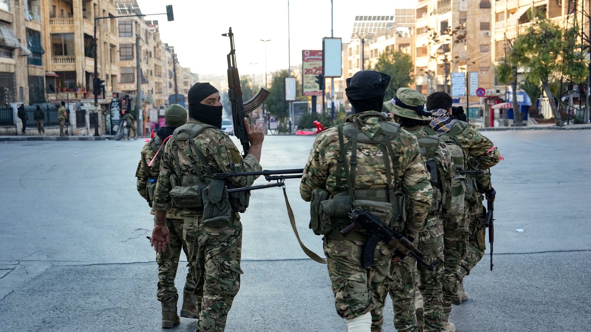 در این تصویر، تعدادی سرباز در یک خیابان urban دیده می‌شود. آن‌ها در حال راه رفتن به سمت دوربین هستند و برخی از آن‌ها سلاح‌های خود را در دست دارند. سربازان لباس‌های نظامی و کلاه‌خودهایی به سر دارند و برخی از آن‌ها صورت‌هایشان را با ماسک پوشانده‌اند. محیط اطراف آنها شامل ساختمان‌ها و خیابان‌های شلوغ است و به نظر می‌رسد در حال گشت‌زنی یا پاسداری از منطقه هستند.