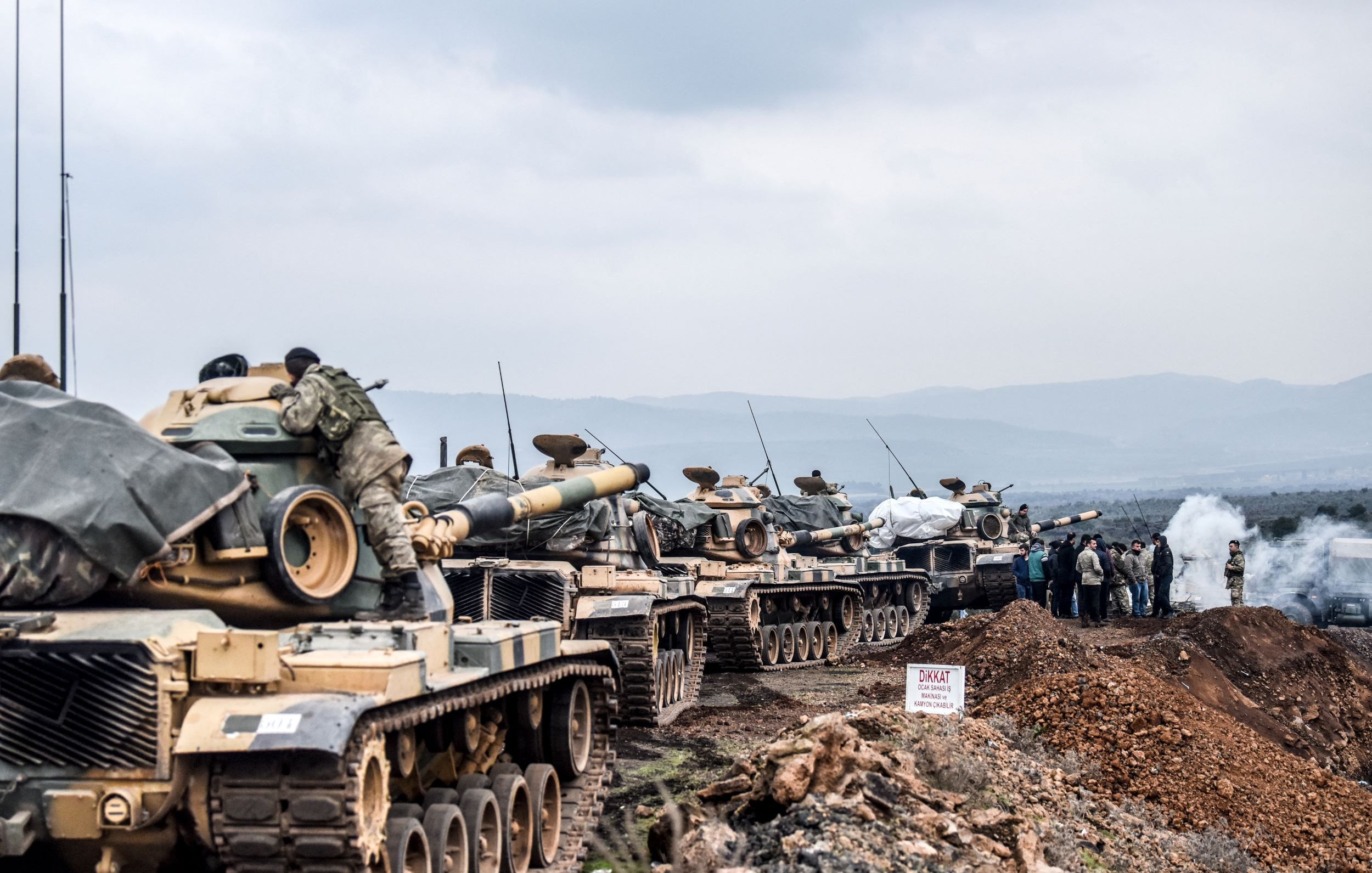 L'image montre une ligne de chars militaires, probablement en position de déploiement ou d'entraînement. Les chars sont recouverts de bâches et semblent être stationnés sur un terrain accidenté, entourés de collines. Au loin, on peut apercevoir des militaires rassemblés autour de ce qui semble être un poste de commandement ou d'observation. Il y a également des signes ou des marquages au sol, et l'atmosphère paraît chargée, avec une légère brume ou fumée dans l'air.