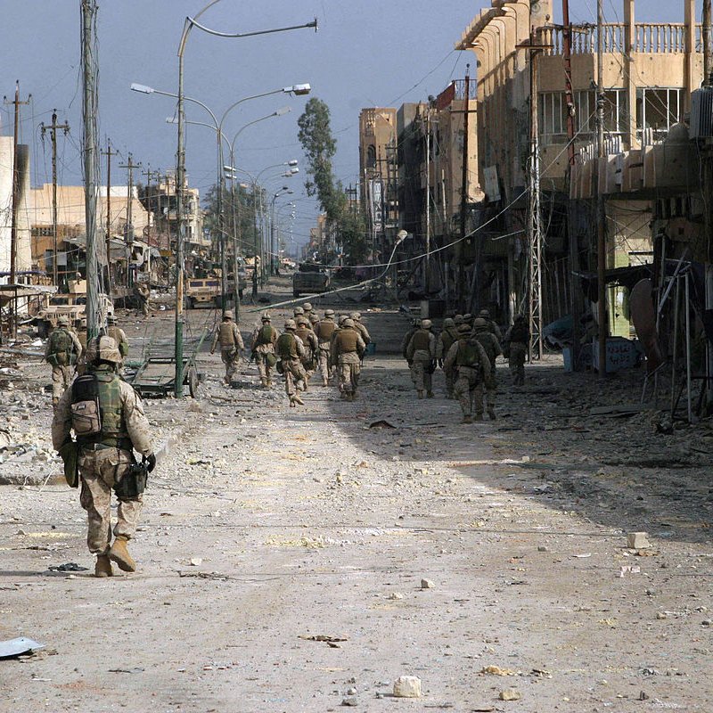 L'image montre une scène urbanisée dévastée, probablement dans une zone de conflit. On y voit des soldats marchant sur une rue déserte, entourés de bâtiments partiellement détruits. Le sol est jonché de débris, et l'atmosphère semble chargée de tension et de désolation. Les structures autour d'eux, avec des fils électriques en désordre, témoignent des ravages causés par le conflit. La lumière est diffuse, ce qui renforce le sentiment d'un environnement difficile et chaotique.