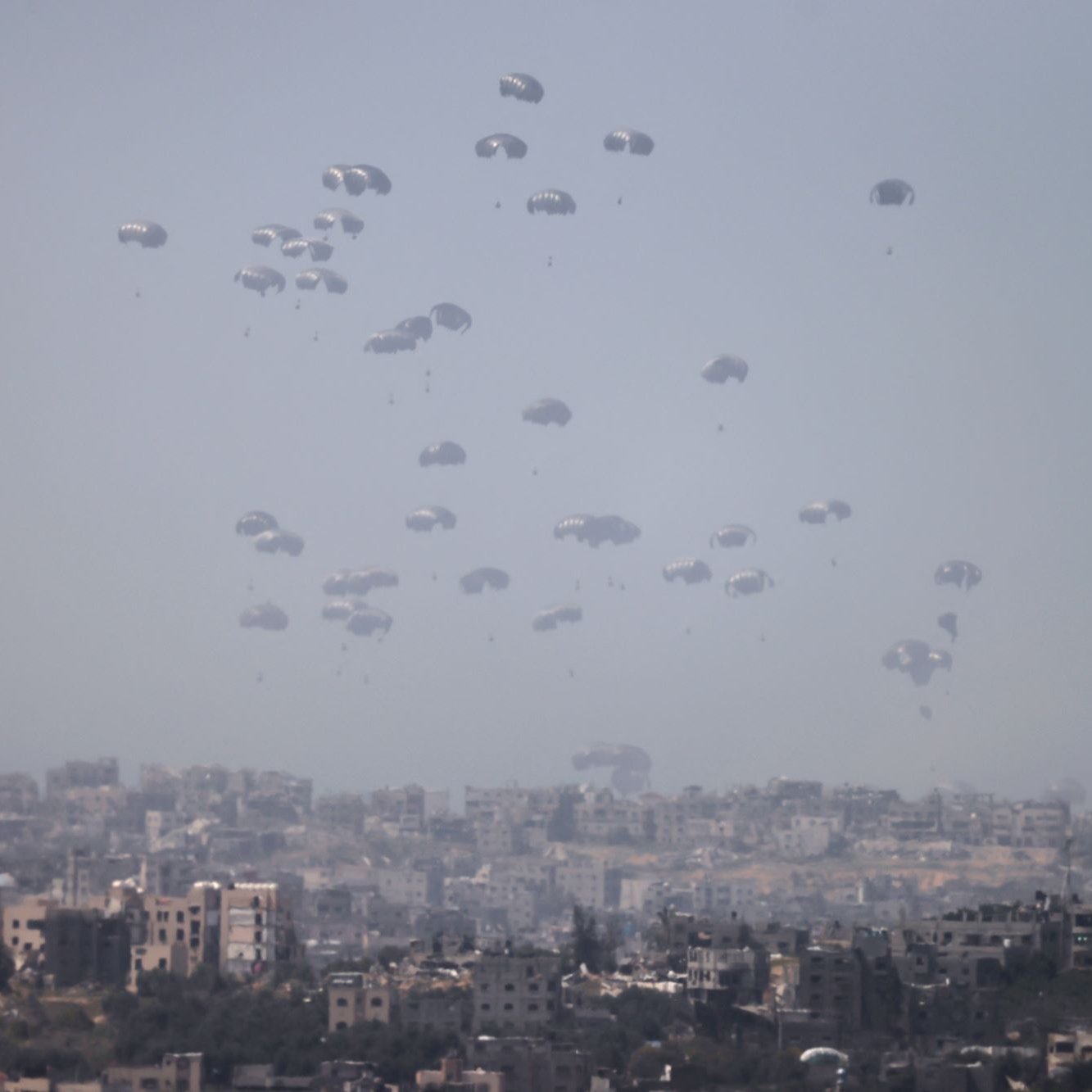 L'immagine mostra un cielo grigio-azzurro con numerosi paracadute che scendono lentamente verso un'area urbana sottostante. Sullo sfondo, è visibile una città con edifici di varie dimensioni e forme. L'atmosfera sembra tesa, suggerendo che si tratta di un'operazione militare o di emergenza. I paracadute, bianchi e leggeri, creano un contrasto visivo con l'ambiente circostante.