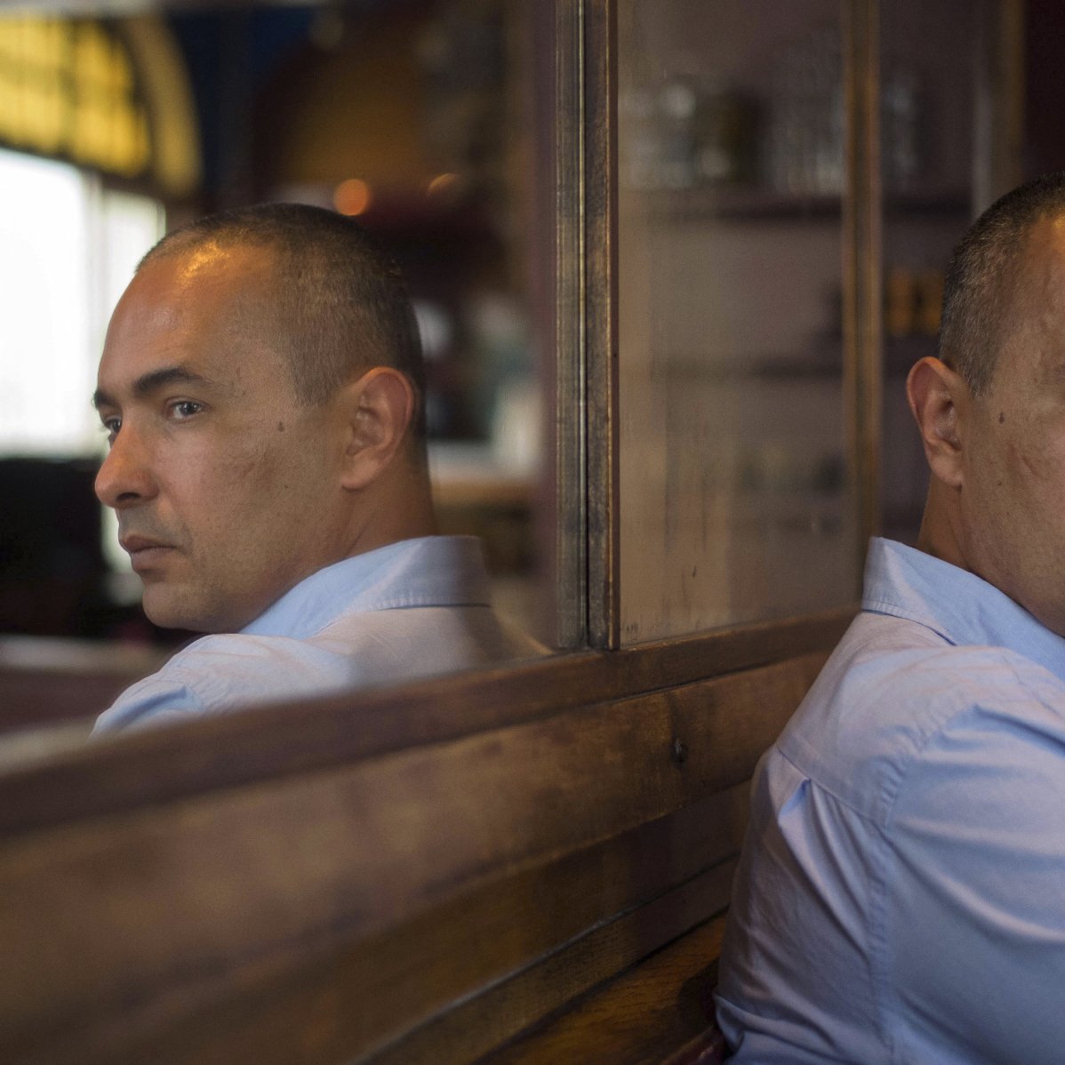 The image features a man sitting with his back to a reflective surface, creating a mirror effect. He has short, dark hair and is wearing a light blue shirt. His expression is pensive, with a serious look on his face as he gazes thoughtfully to the side. The surroundings indicate a cozy indoor setting, with warm lighting and wooden elements in the background. The reflection captures both his profile and the contemplative mood of the scene.