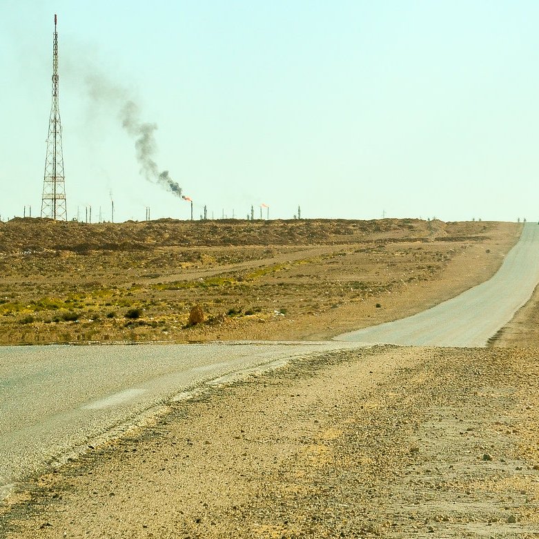 L'image montre un paysage désertique avec une route qui s'étend à perte de vue. À droite, un panneau indiquant "Hassi R'Mel" avec des chiffres. En arrière-plan, on aperçoit une tour de communication et une colonne de fumée émanant d'une installation, probablement liée à l'industrie pétrolière. Le sol est aride, et il n'y a pas beaucoup de végétation environnante, illustrant un environnement rural et isolé.