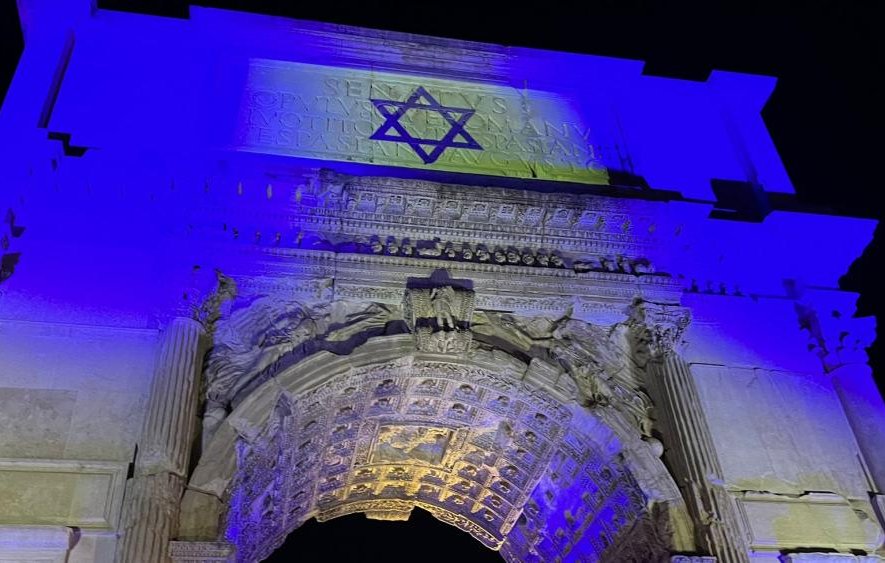 L'immagine mostra un arco a un'altezza notevole, illuminato da luci blu. Sulla parte superiore dell'arco è presente una stella di David, simbolo del giudaismo. L'architettura è riccamente decorata con dettagli ornamentali, evidenziando colonne e incisioni. L'ambientazione notturna conferisce all'opera una presenza suggestiva e solenne.