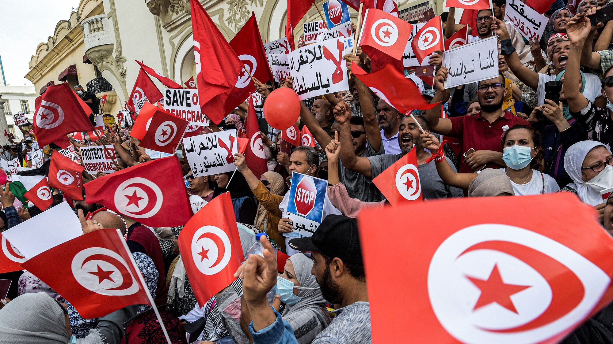 L'image montre une foule manifestante en Tunisie, brandissant des drapeaux tunisiens et des pancartes. Les participants expriment des messages en faveur de la démocratie, avec des slogans tels que "Sauvez la démocratie" et "Stop". On peut voir des gens de différents âges et genres, certains portant des masques. L'ambiance semble énergique et engagée, avec un fort esprit de revendication collective.