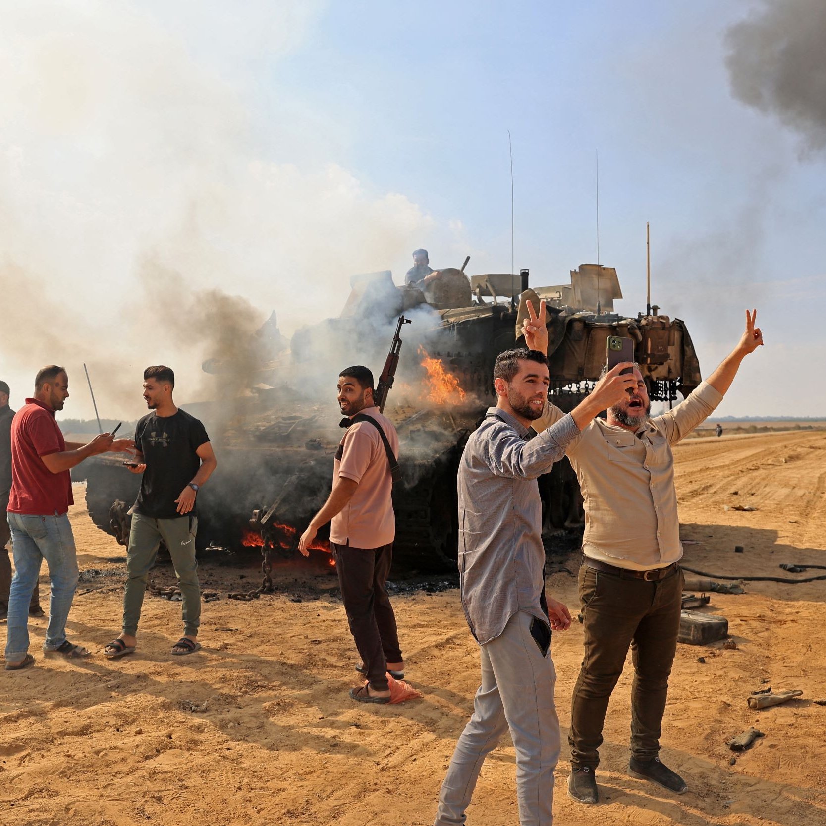 L'immagine mostra un gruppo di persone in un ambiente di conflitto. In primo piano, alcuni uomini stanno facendo gesti di vittoria e sembrano esprimere emozioni forti. Sullo sfondo, si vede un veicolo distrutto, avvolto da fumi e fiamme. Il terreno appare sabbioso e polveroso, e ci sono nuvole di fumo nero che si alzano verso il cielo. L'atmosfera è intensa e suggerisce un contesto di tensione e conflitto.