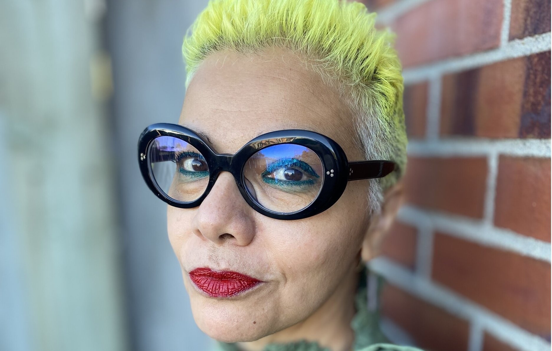 L'immagine mostra una persona con capelli corti e di colore giallo brillante. Indossa occhiali grandi e neri, che aggiungono un tocco audace al suo look. Il trucco è accentuato con labbra rosse, mentre lo sfondo presenta una parete di mattoni, creando un contrasto interessante con i colori vivaci della persona. La sua espressione è sicura e affermativa.