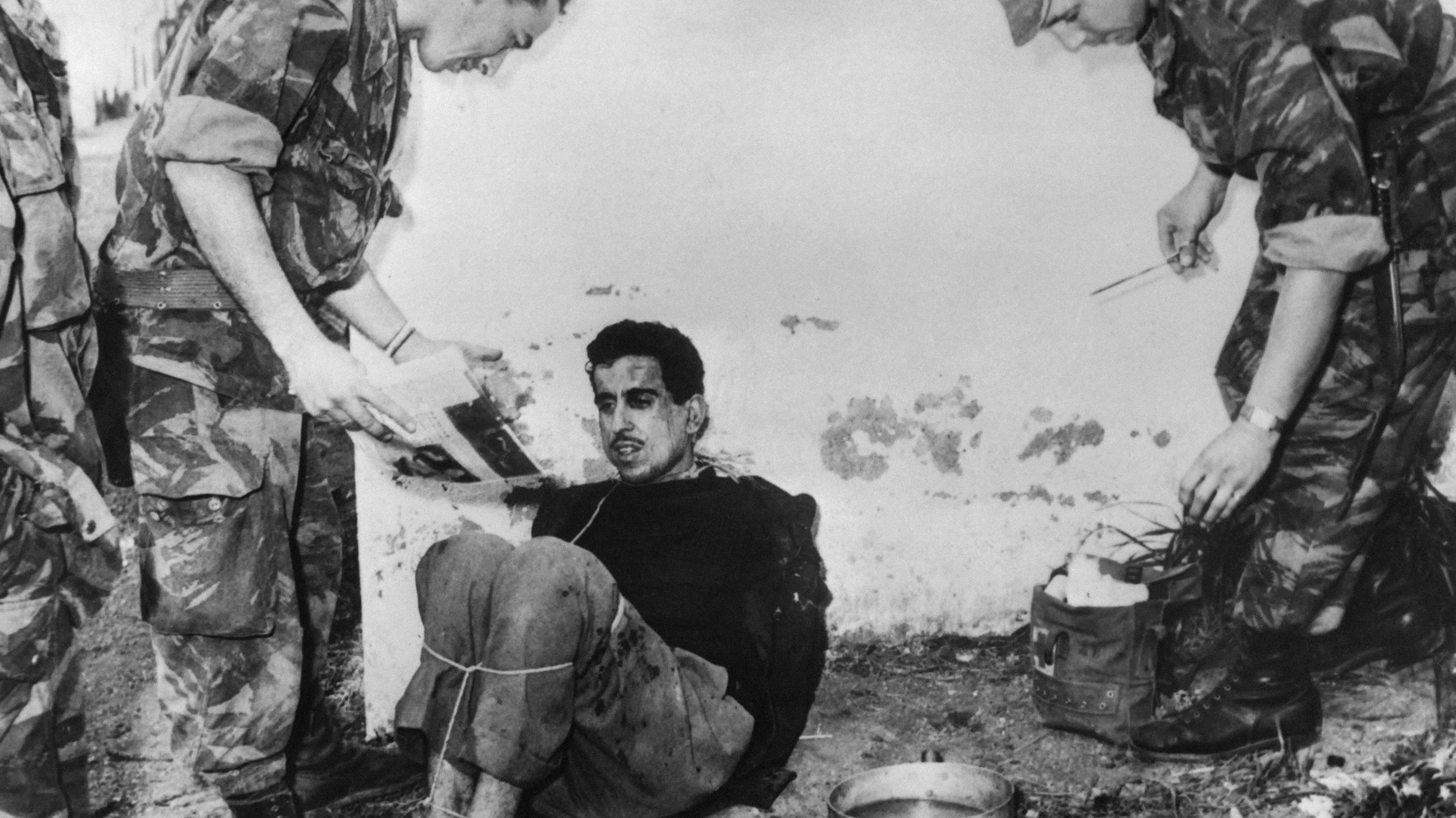 La imagen muestra a un hombre sentado en el suelo, atado y aparentemente en una situación de interrogatorio o detención. Tiene una expresión de tensión en su rostro. Dos personas, vestidas con uniformes militares, están de pie a su alrededor. Una de ellas sostiene un documento y parece que está hablando con el detenido, mientras que la otra sostiene un utensilio, probablemente para tomar notas o hacer preguntas. El ambiente es austero y parece un lugar al aire libre, con una pared desgastada de fondo. La escena capta un momento de tensión y conflicto.