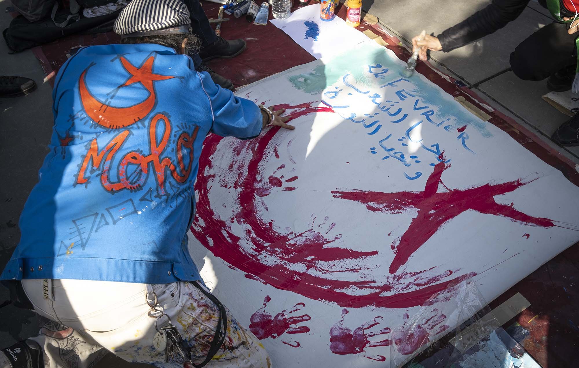 L'image montre une personne en train de peindre sur une grande toile au sol. La personne porte une veste bleu avec le mot "Moh" écrit dessus. On peut voir des éclaboussures de peinture rouge et verte, et un motif en forme de croissant de lune et d'étoile, qui sont des symboles souvent associés à certains pays. Des inscriptions sont également visibles sur la toile. L'environnement semble être celui d'une manifestation ou d'un évènement artistique.