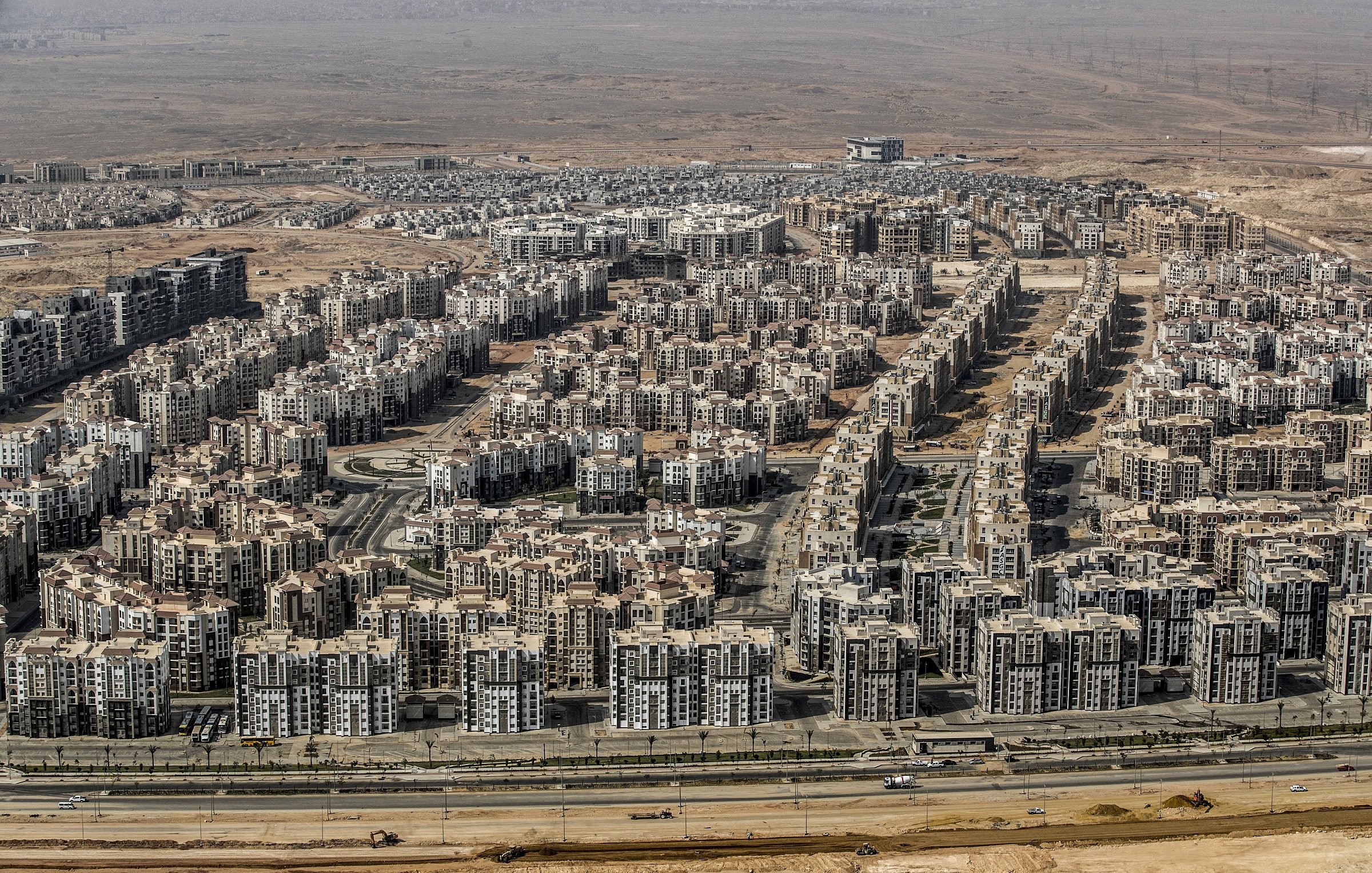 L'image montre une vaste zone urbaine composée de nombreux immeubles de plusieurs étages, organisés de manière dense. On peut voir des routes qui traversent ces secteurs résidentiels, avec des infrastructures modernes. En arrière-plan, le paysage est désertique, ce qui évoque un contraste entre l'urbanisation et l'environnement naturel environnant. L'ensemble dégage une impression de développement urbain important dans une région aride.