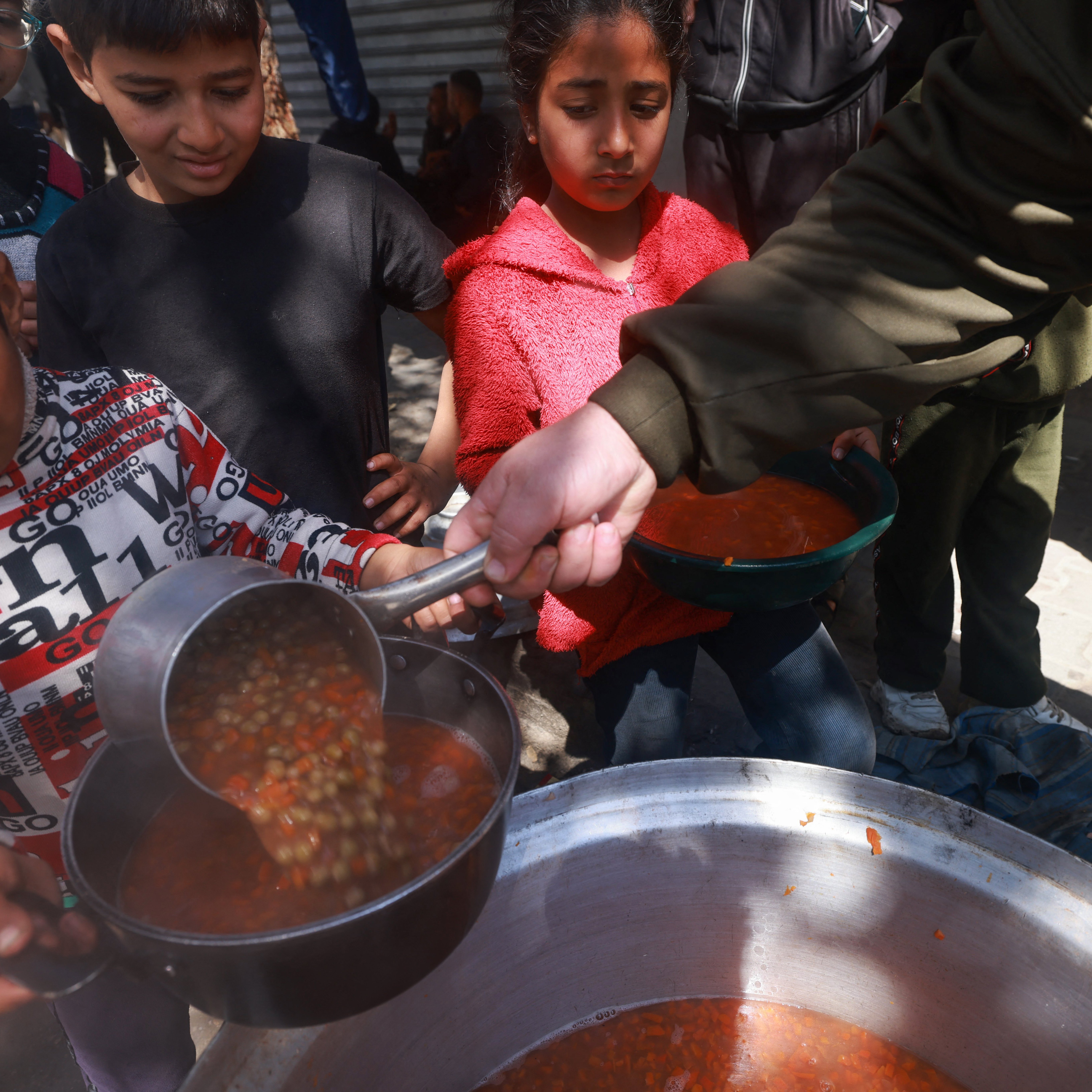 L'immagine mostra un gruppo di bambini in un ambiente all'aperto, mentre qualcuno sta servendo del cibo da una grande pentola. Un bambino, in particolare, sta utilizzando un mestolo per prendere una porzione di una zuppa o stufato, che sembra contenere legumi. Gli altri bambini attendono, con espressioni curiose e attente, alcuni con uno sguardo serio. L'atmosfera trasmette un senso di comunità e condivisione, evidenziando un momento significativo di aiuto e sostegno.