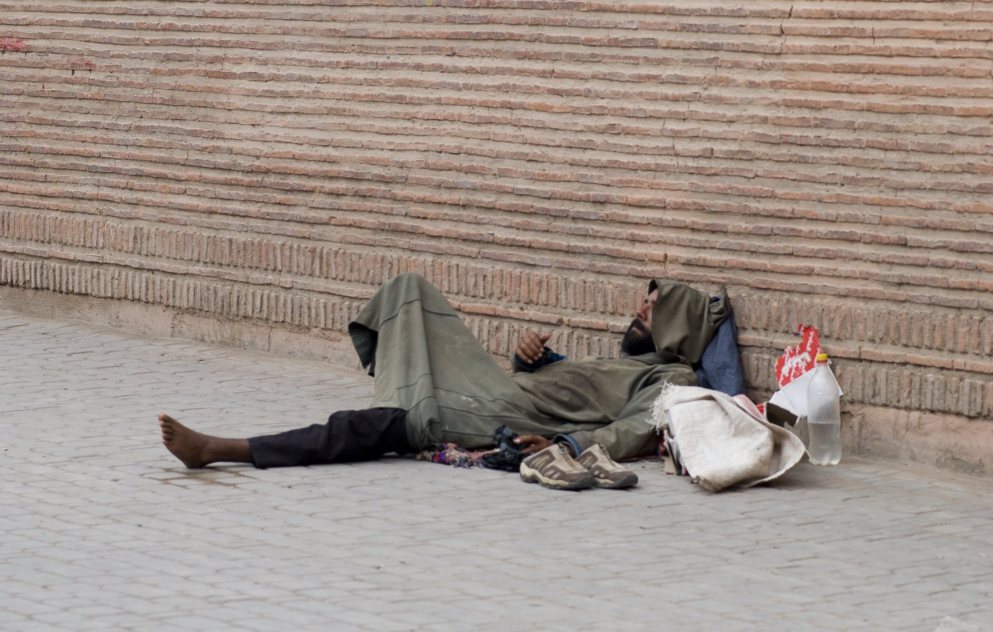 L'image montre une personne allongée sur le sol, semblant sans abri. Elle porte des vêtements sombres et usés, et se trouve à côté d'une paroi de briques. On voit des objets éparpillés autour d'elle, comme des chaussures et un sac. L'environnement semble urbain et le ton de l'image est contemplatif.