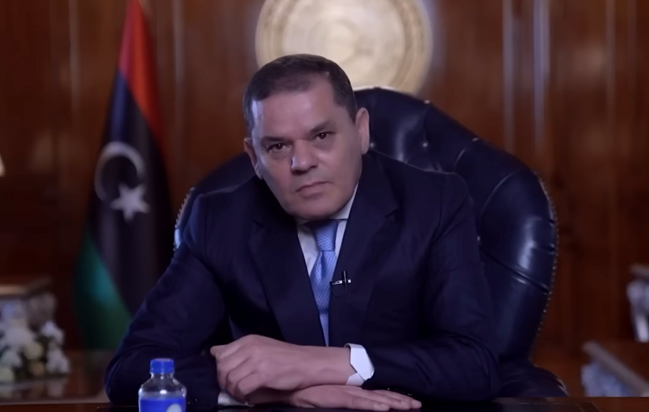 Homme en costume assis dans un bureau, drapeau libyen en arrière-plan.