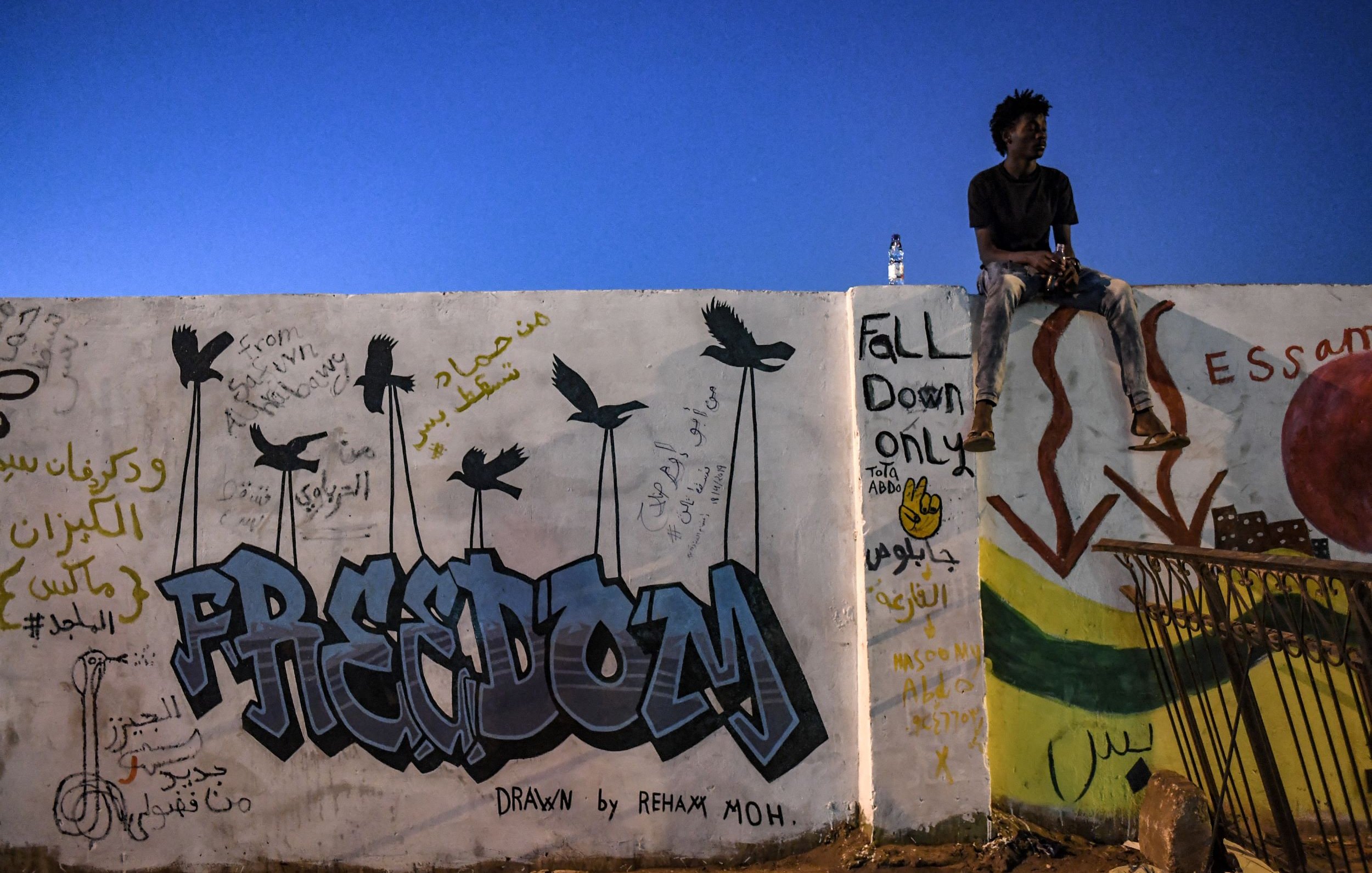 L'image montre un mur couvert de graffitis avec le mot "FREEDOM" en lettres colorées. À côté, des dessins d'oiseaux sont visibles. Un jeune homme est assis sur le bord du mur, regardant au loin, tandis qu'une bouteille d'eau est posée à côté de lui. Le ciel est d'un bleu profond, indiquant une prise de vue en soirée. Des inscriptions en arabe et des dessins supplémentaires peuvent être observés sur le mur.