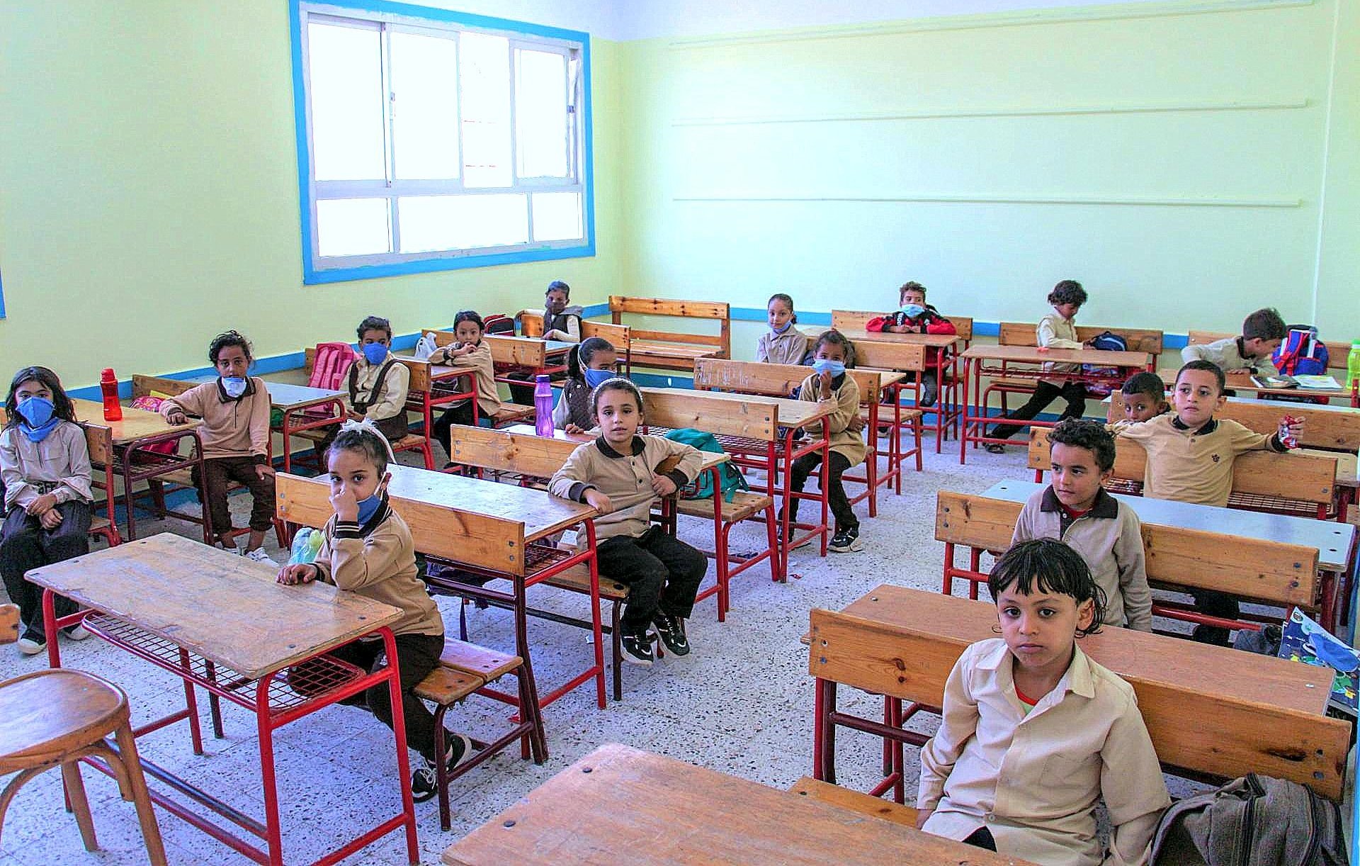 L'image montre une salle de classe avec plusieurs élèves assis à des bureaux en bois. Les enfants portent des uniformes beige et plusieurs d'entre eux ont des masques sur le visage. La pièce est lumineuse, avec des murs peints en jaune clair et des fenêtres qui laissent entrer la lumière. Les élèves semblent attentifs, certains regardant vers l'appareil photo. Les bureaux sont espacés, et il y a quelques fournitures scolaires visibles sur les tables.