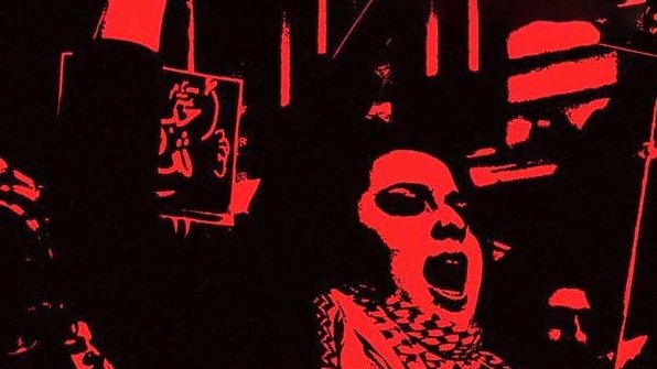 L'image présente une scène expressive et énergique, probablement de manifestation ou de rassemblement. On y voit une personne au premier plan qui lève son bras, suggérant un appel à l'action ou une déclaration de protestation. L'arrière-plan est dominé par des couleurs sombres, ce qui intensifie le sentiment de détermination. Des affiches ou des slogans en arabe sont visibles, renforçant l'idée d'un message collectif. L'atmosphère évoque un fort engagement et une passion pour une cause.