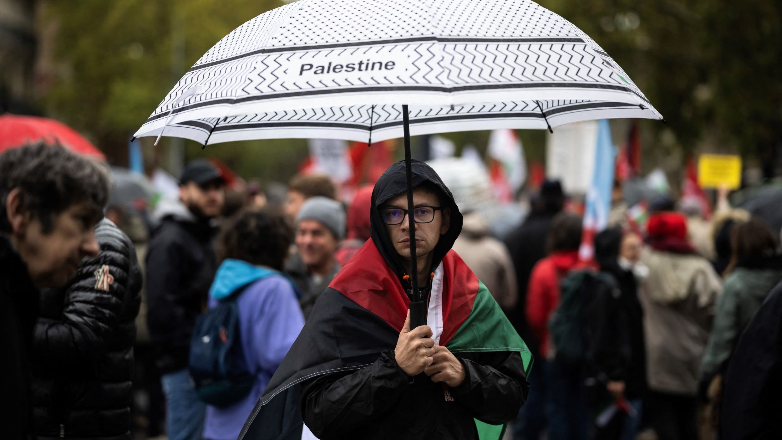 En la imagen, se puede ver a una persona que sostiene un paraguas con la palabra "Palestine" escrita en él. La persona lleva una bandera de Palestina sobre los hombros y parece estar en una manifestación o protesta. Alrededor se pueden observar otras personas, algunas con paraguas y banderas, en un ambiente cargado de emociones y solidaridad. El clima parece nublado o lluvioso, lo que añade a la atmósfera del evento.