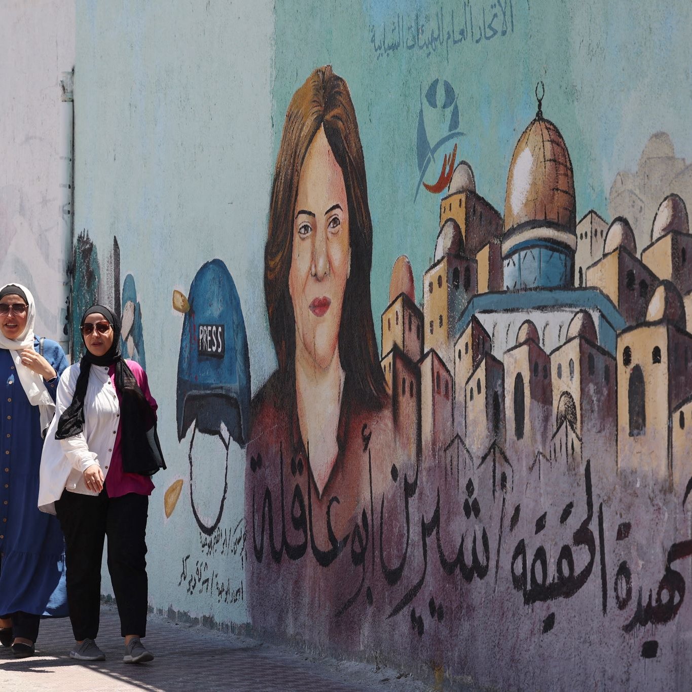 L'image montre deux femmes marchant le long d'un mur décoré d'une fresque. La fresque représente le portrait d'une femme avec un casque portant l'inscription "PRESS", suggérant qu'elle est journaliste. En arrière-plan, on aperçoit des maisons stylisées qui pourraient évoquer une ville ou un quartier particulier. Le texte en arabe est également visible, mais seul le contexte artistique et le message de solidarité ou de mémoire peuvent être interprétés. L'atmosphère est animée et engagée.
