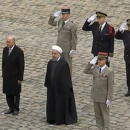 L'image montre plusieurs personnes en uniforme militaire saluant. Au centre, deux hommes en costume : l'un à gauche semble être un dirigeant, tandis que l'autre, portant une robe noire et un turban, est probablement un haut responsable religieux ou politique. Les autres personnes en arrière-plan sont des officiers en uniforme, également en train de rendre les honneurs. Ils se trouvent sur une surface en pavés, suggérant une cérémonie officielle ou un événement diplomatique.