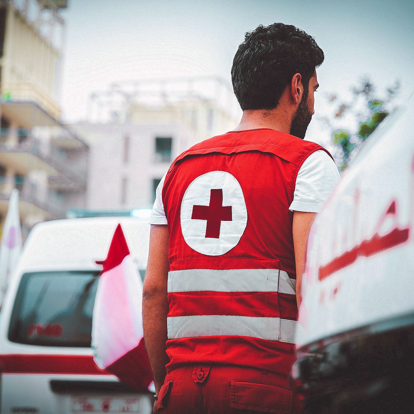 الصورة تظهر شابًا يرتدي زيًا خاصًا بالهلال الأحمر، والذي يتكون من سترة حمراء مزينة بعلامة الصليب الأحمر. يبدو الشاب من الخلف، وهو يقف بالقرب من سيارات إسعاف تحمل شعار الهلال الأحمر. تظهر في الخلفية مباني ومرافق عامة، مما يشير إلى وجود أنشطة أو أحداث إنسانية في المنطقة. تعكس الصورة الجهد الإنساني ورغبة المساعدة في الحالات الطارئة.