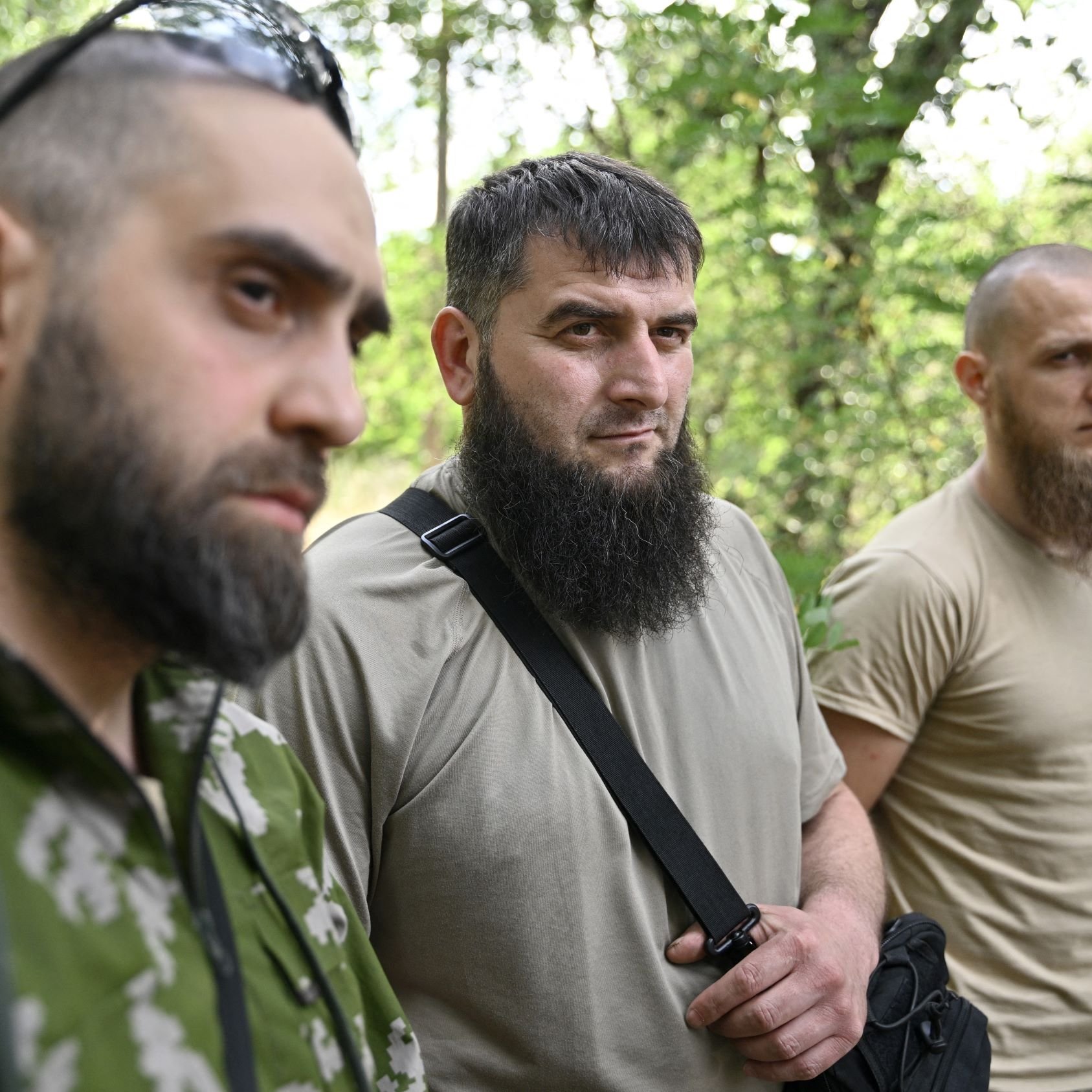 L'image montre trois hommes se tenant côte à côte dans une forêt. Tous trois portent des vêtements utilitaires, et chacun d'eux a une barbe. Leurs expressions sont sérieuses, et l'éclairage naturel met en valeur leur présence dans ce cadre boisé. On peut observer des arbres en arrière-plan, ce qui crée une ambiance naturelle.
