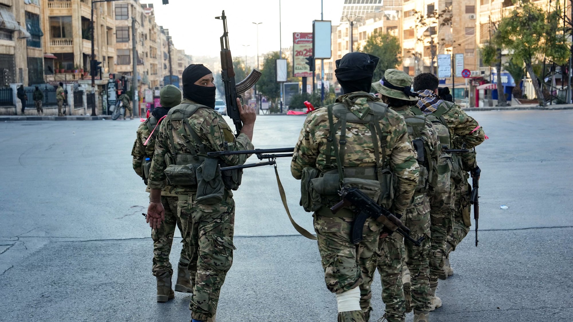 L'immagine mostra un gruppo di soldati in abbigliamento militare che camminano in una strada urbana. I soldati sono armati e indossano maschere che coprono parte del volto. Sullo sfondo si possono vedere edifici e un ambiente urbano, con luci e segnali stradali visibili. L'atmosfera è quella di un contesto di sicurezza o di operazioni militari.