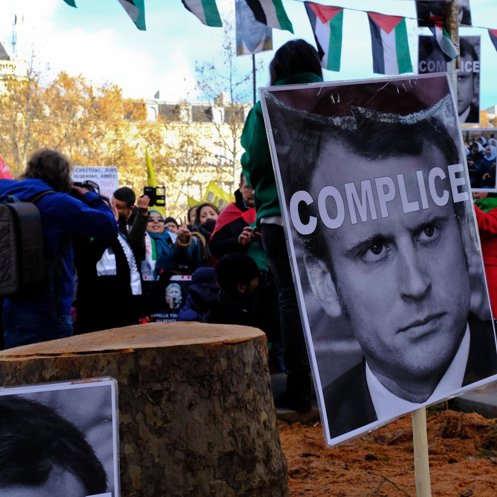 L'image montre une manifestation où une foule se rassemble autour d'un tronc d'arbre coupé. Des panneaux sont affichés avec des portraits et le mot "COMPLICE" écrit en gros caractères sur une affiche. En arrière-plan, on peut voir des drapeaux et des personnes prenant part à la protestation. L'atmosphère semble tendue, illustrant des sentiments de mécontentement et des revendications politiques.