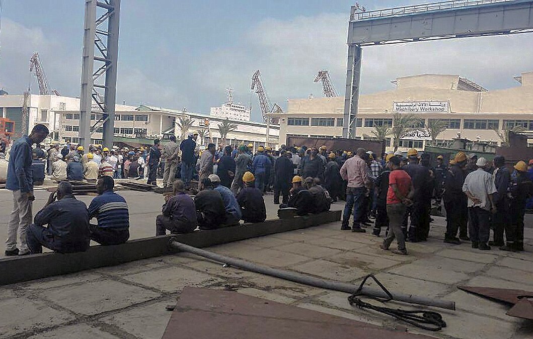 L'image montre un groupe de nombreux travailleurs rassemblés dans un espace extérieur, probablement dans un port ou un chantier naval. Les personnes sont vêtues de tenues de travail et portent des casques de sécurité jaunes. Certains d'entre eux sont assis sur le sol, tandis que d'autres sont debout, discutant ou attendant. L'environnement semble industriel, avec des structures métalliques et des équipements visibles en arrière-plan. Le ciel est partiellement nuageux.