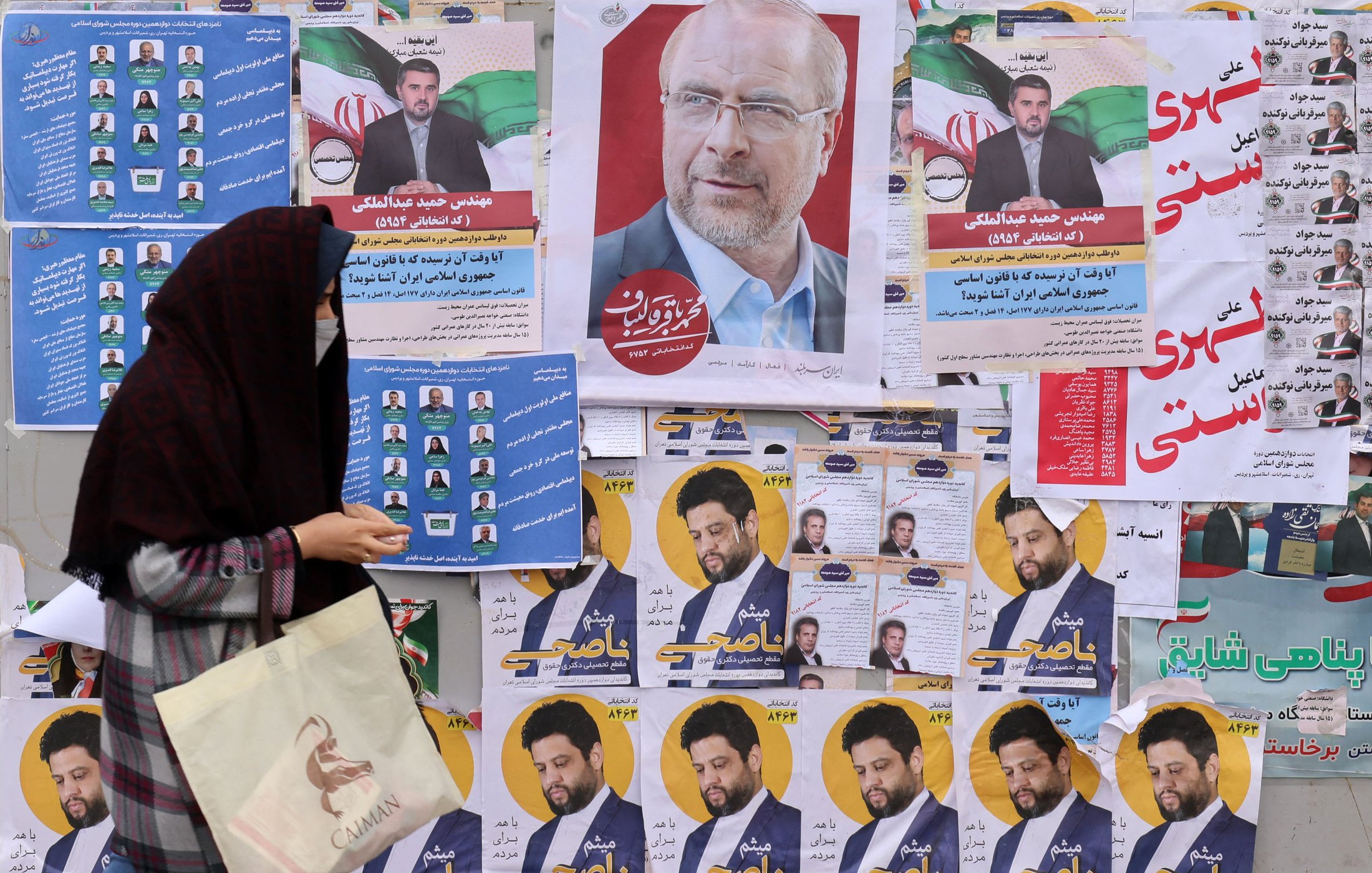 L'image montre un mur recouvert de diverses affiches électorales. On peut voir plusieurs portraits de candidats, accompagnés de slogans et d'informations. Au premier plan, une femme portant un hijab et tenant un sac semble marcher devant ces affiches, ce qui suggère une ambiance électorale. Les couleurs des affiches sont vives et variées, ajoutant à l'aspect animé de la scène.