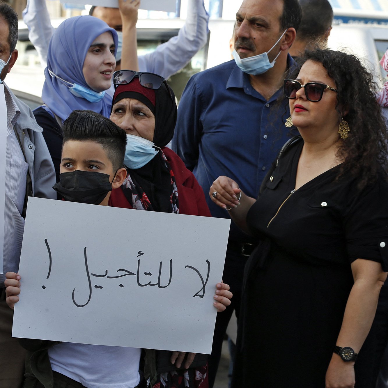 The image depicts a group of people participating in a protest. Some individuals are wearing masks, and they are holding signs, including one that prominently features the Arabic phrase "لا للتأجيل" (translated as "No to delay"). The crowd appears to include men, women, and children, with a mix of facial expressions that suggest determination and solidarity. The background shows a public space, likely in an urban setting.