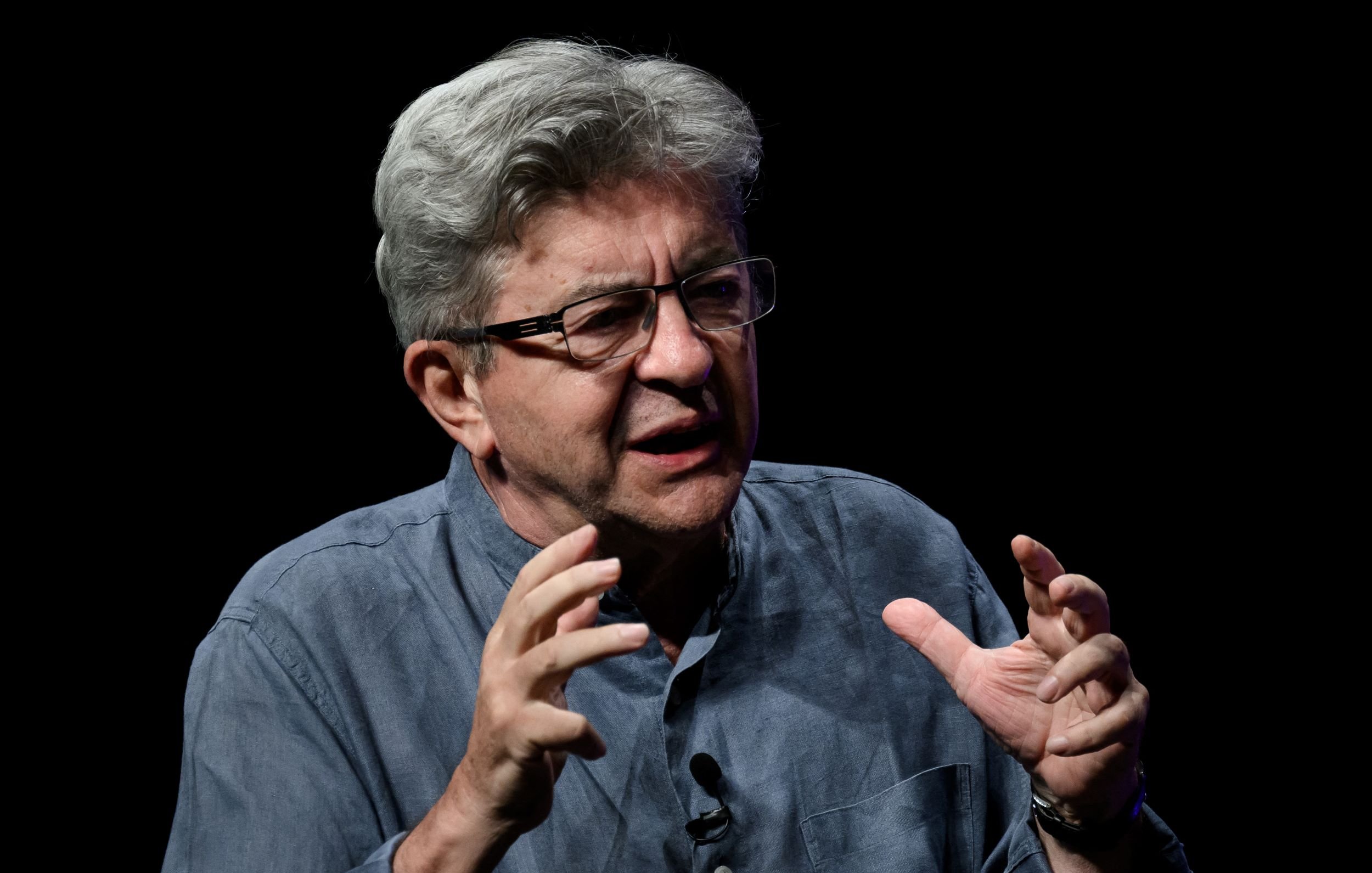 في الصورة، يظهر رجل مسن ذو شعر رمادي وملامح تعبر عن الحماس أو التركيز. يرتدي قميصًا رماديًا ويبدو أنه يتحدث بشكل نشط، حيث يستخدم يديه للتعبير عن أفكاره. الخلفية مظلمة، مما يجعل التركيز بالكامل على الشخص الذي يتحدث. تعكس تعابير وجهه والشكل الذي يتحدث فيه شغفًا أو حماسًا لموضوعه.