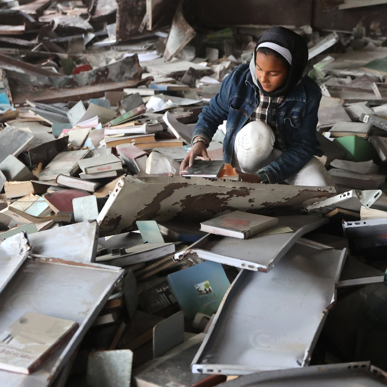 تصویر یک اتاق شلوغ و پر از کتاب‌های پراکنده است. یک دختر جوان در حال نشسته بر روی زمین، در حال بررسی یا جستجوی چیزی در میان کتاب‌ها و وسایل دیگر است. او لباس‌های ساده‌ای به تن دارد و فضای اطرافش بی‌نظم و به هم ریخته به نظر می‌رسد.