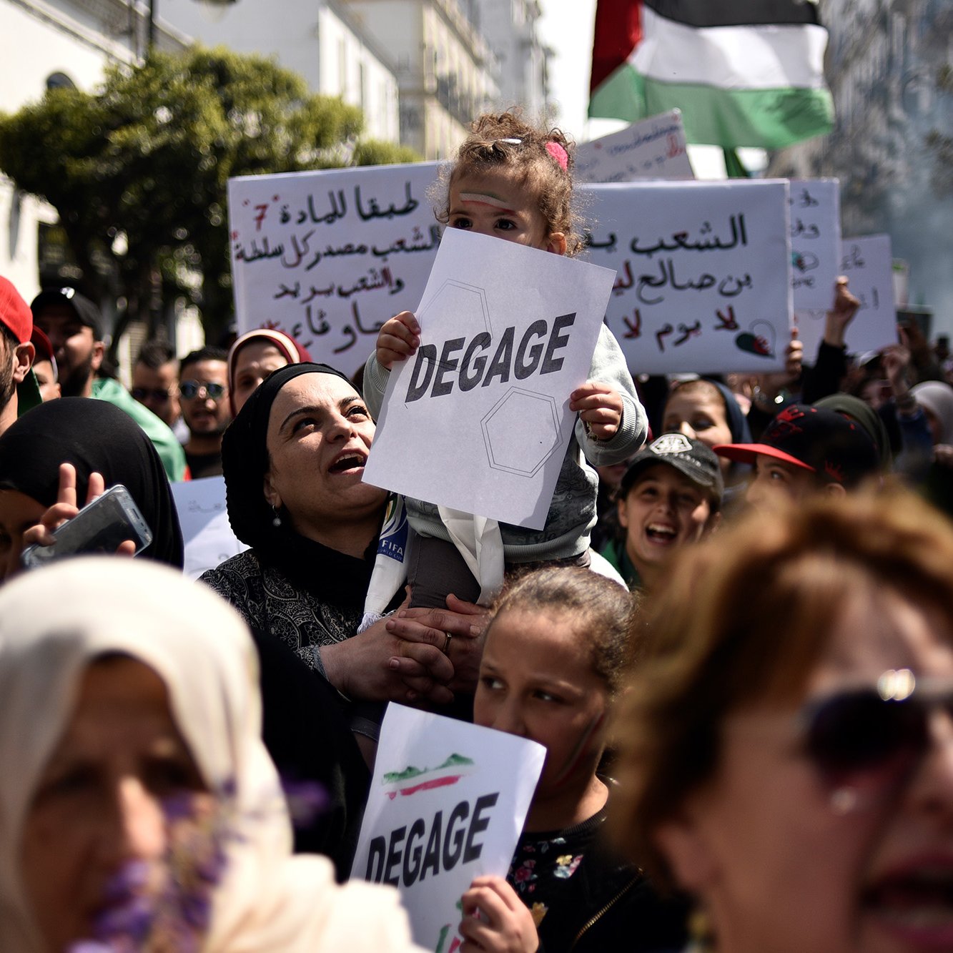 L'image montre une foule rassemblée pour une manifestation. Au premier plan, une femme porte un enfant sur ses épaules, et elle tient un panneau avec le mot "DEGAGE". Les manifestants semblent exprimer des revendications politiques. On peut également voir d'autres panneaux en arabe et des drapeaux, ce qui indique une atmosphère de protestation collective. La scène se déroule en plein air, probablement dans une ville, et les expressions des participants montrent une forte détermination.