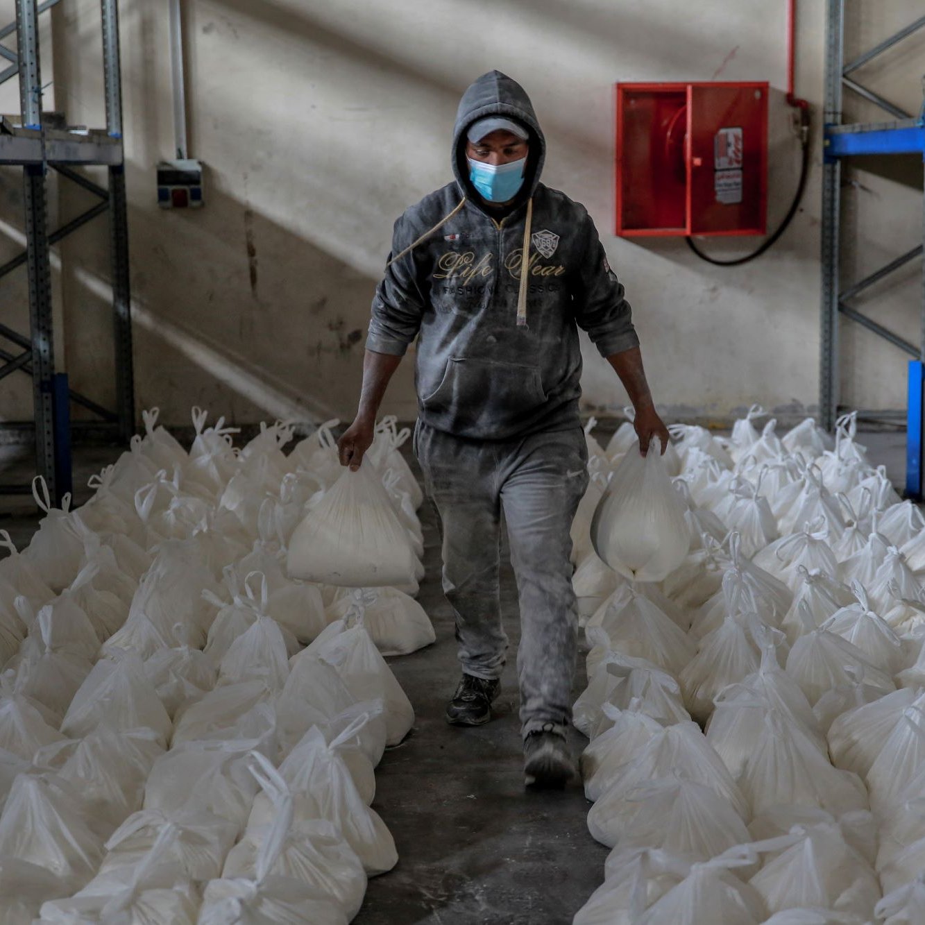Sur cette image, on voit une personne portant un sweat à capuche gris et un masque de protection. Elle marche à travers une pièce de stockage ou un entrepôt, tenant deux sacs blancs en plastique. De nombreux sacs similaires sont disposés de manière ordonnée sur le sol autour de la personne. Les murs de la salle semblent ternes et l'éclairage suggère un environnement de travail modeste. Au fond, on peut voir une boîte de protection contre les incendies fixée au mur.