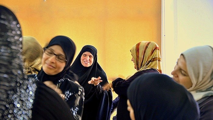 L'image montre un groupe de femmes en discussion ou en interaction. Elles portent des vêtements traditionnels, y compris des hijabs et des abayas. Certaines femmes sourient et semblent engagées dans une conversation joyeuse. L'éclairage dans la pièce donne une ambiance chaleureuse, avec un fond qui semble jaune. L'atmosphère générale est conviviale et animée.