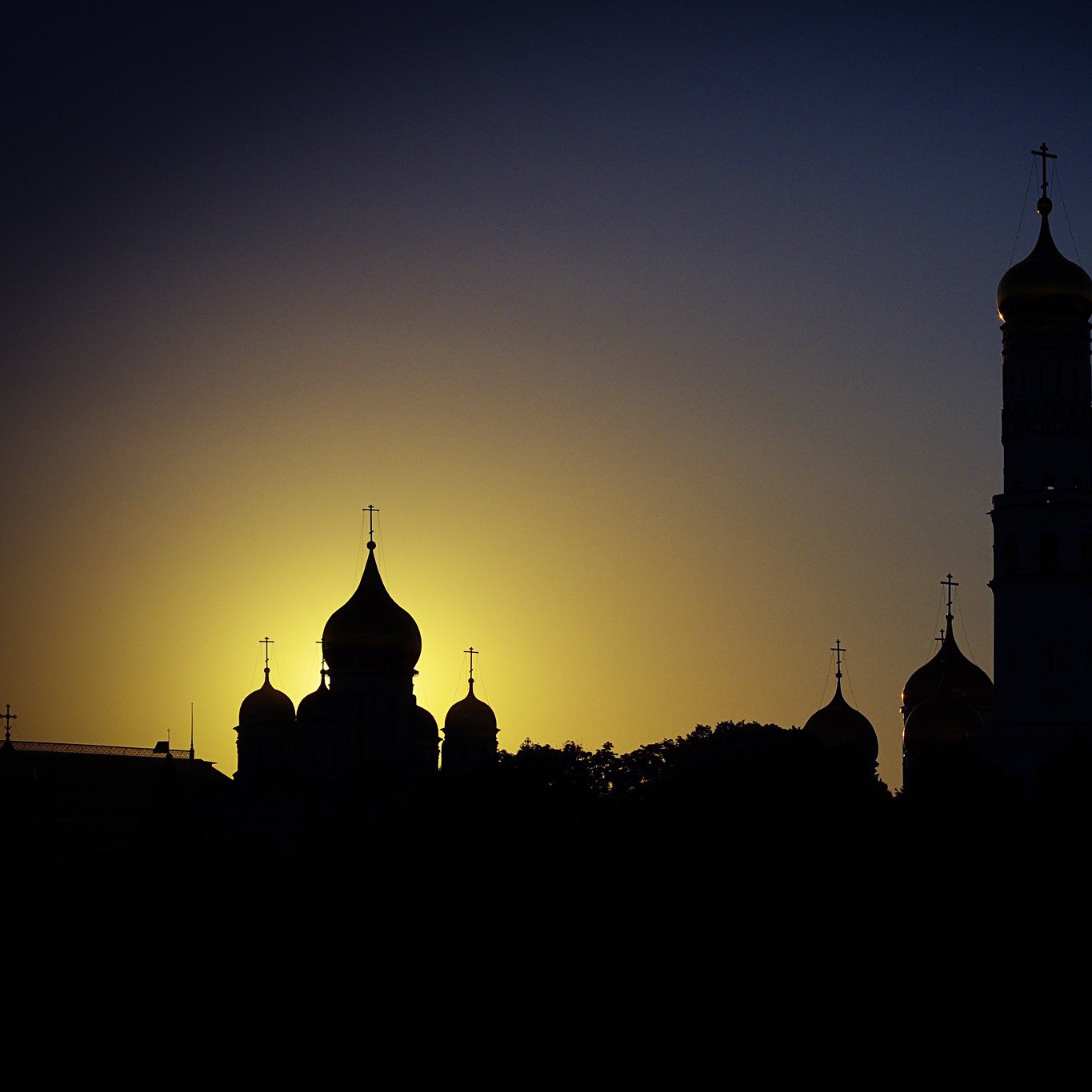 تظهر في الصورة silhouettes لعدد من الكنائس مع قبابها وأبراجها في لحظة غروب الشمس. الخلفية تميل إلى الألوان الدافئة للغروب، مع تدرجات من الأصفر إلى الأزرق الداكن. تبرز المعالم المعمارية بشكل جميل ضد السماء، مما يضفي على الصورة جوًا من الهدوء والجمال الروحي.