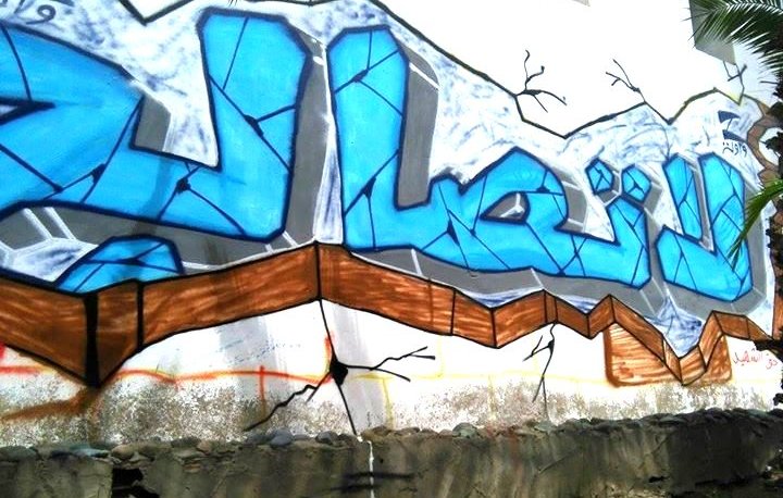 L'image montre un mur de graffiti avec des lettres stylisées en bleu. Le fond est légèrement cassé, ce qui donne l'impression que le mur est en train de se fissurer. Le mot écrit semble être en arabe, avec des motifs et des contours en noir qui accentuent le design. En bas, on peut apercevoir des déchets et de l'herbe, ce qui montre que l'art se trouve dans un environnement urbain.