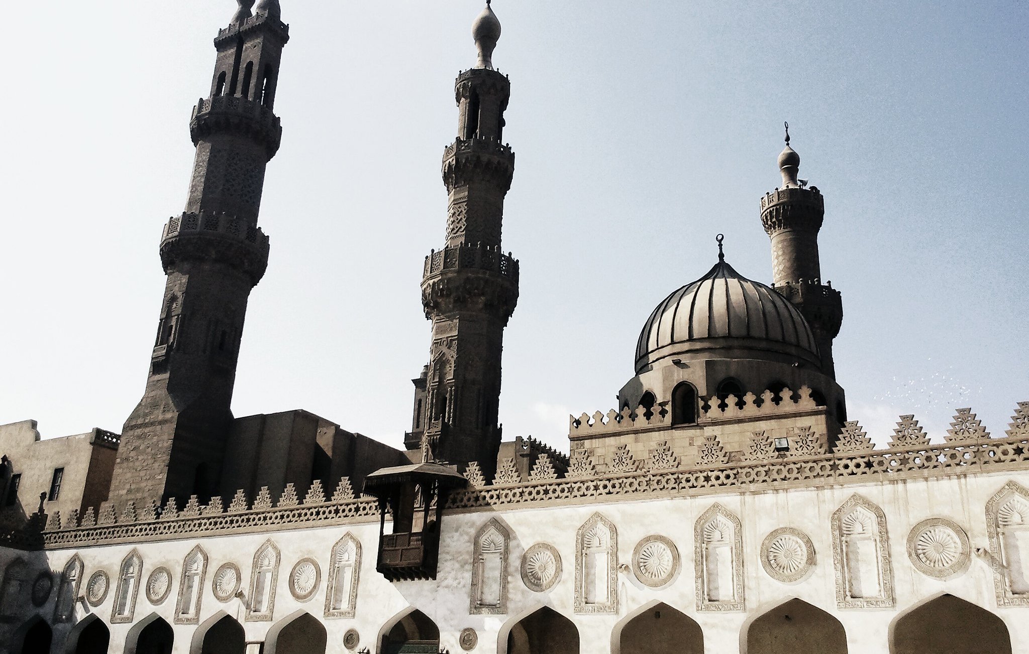 L'image montre une mosquée majestueuse avec deux minarets qui s'élèvent haut dans le ciel. La structure présente une belle architecture islamique, caractérisée par des arches décoratives et des motifs ornementaux sur les murs. Le dôme au centre ajoute à la grandeur du bâtiment, tandis que le ciel clair en arrière-plan donne une ambiance lumineuse à la scène. Les détails sculptés et la symétrie des éléments architecturaux témoignent d'un riche héritage culturel.