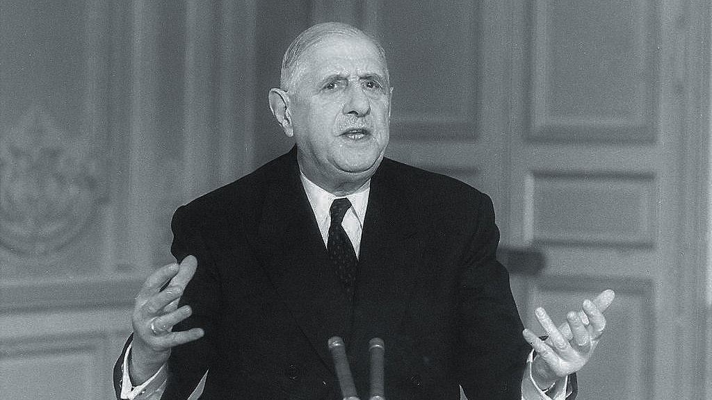 The image depicts a man in a formal suit seated at a desk, speaking with his hands raised expressively. He appears to be delivering a speech or addressing an audience. The setting suggests a formal environment, likely a governmental or political space, with a plain background that emphasizes the speaker. The monochromatic tone gives the image a historical feel, suggesting it may be from a past era.