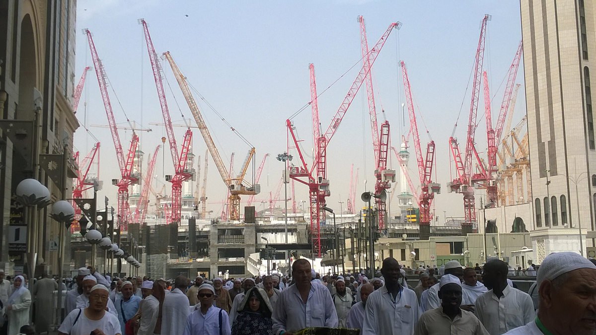 L'image montre un grand nombre de personnes, probablement des pèlerins, se déplaçant dans une zone de construction. En arrière-plan, on peut voir de nombreuses grues rouges qui soulignent les travaux en cours. Les pèlerins portent des vêtements traditionnels, souvent blancs, ce qui est courant lors des pèlerinages. L'environnement semble animé, avec une ambiance de mouvement et d'activité en raison des constructions.