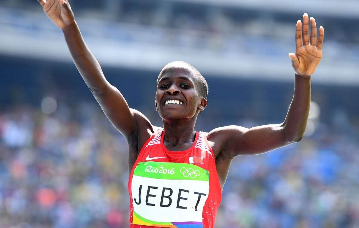 L'image montre une athlète en plein moment de célébration. Elle a les bras levés en l'air, affichant un large sourire, ce qui suggère qu'elle vient de réaliser une performance exceptionnelle. Elle porte un maillot rouge avec son nom "JEBET" inscrit dessus, ainsi que des éléments des Jeux Olympiques de Rio 2016. L'arrière-plan montre une foule et un stade, indiquant qu'elle est en compétition dans un événement majeur.