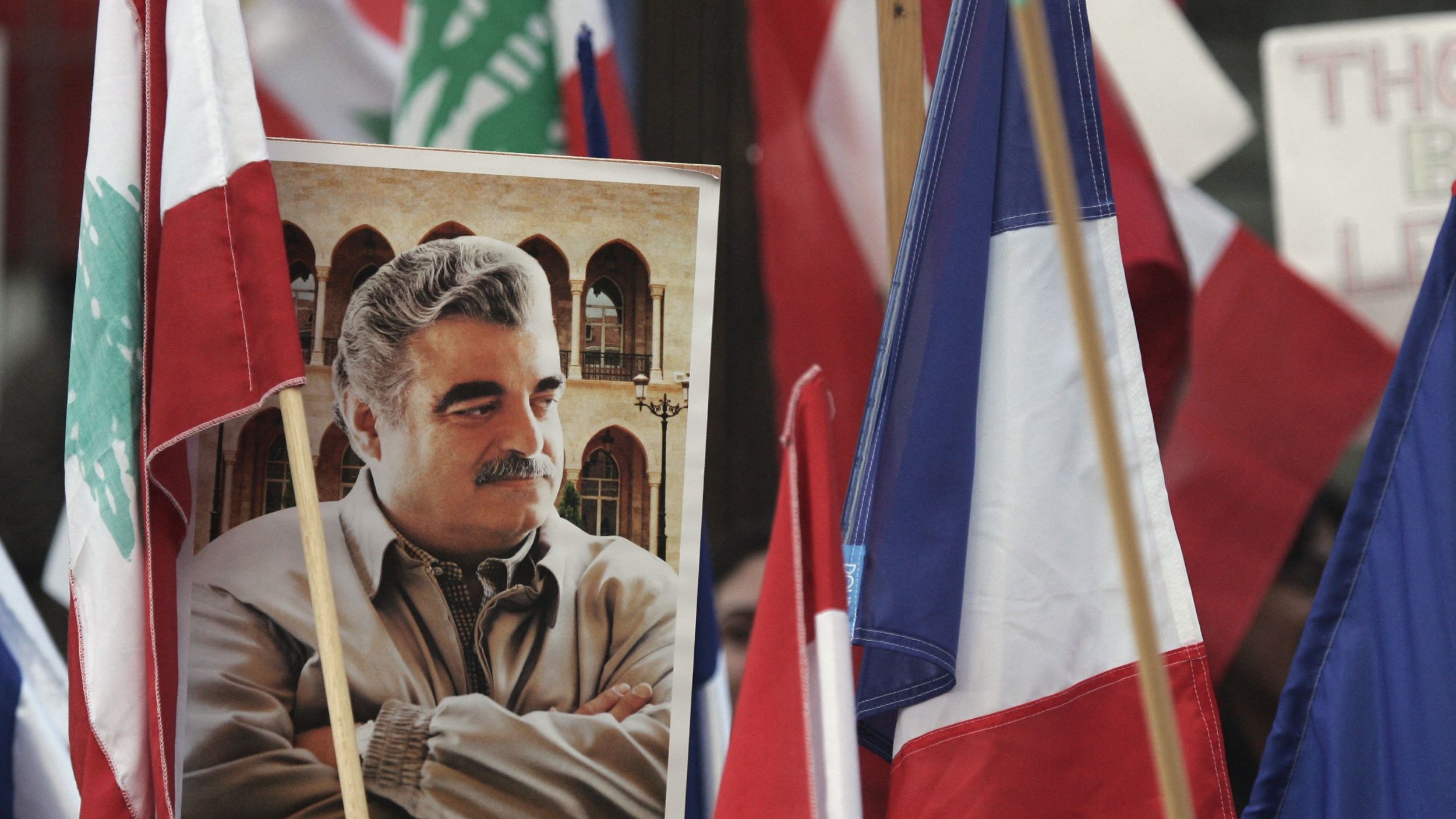 تظهر في هذه الصورة مجموعة من الأعلام، بما في ذلك العلم اللبناني والعلم الفرنسي. في وسط الصورة، هناك صورة كبيرة لرجل يبدو أنه شخصية معروفة، حيث يظهر واقفًا مع ذراعيه متقاطعتين. الخلفية تضم مجموعة من الأشخاص الذين يحملون الأعلام، مما يدل على أن هذه الصورة قد التقطت خلال فعالية أو تجمع يمثل قضية معينة.