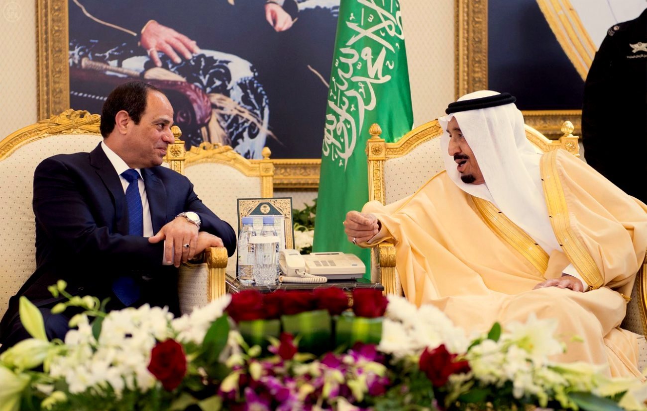 L'image montre deux hommes assis face à face dans un cadre officiel. Ils sont vêtus de manière formelle : l'un porte un costume noir avec une cravate, tandis que l'autre est en tenue traditionnelle dorée. Ils sont entourés d'une décoration luxueuse, avec des drapeaux en arrière-plan et des fleurs sur une table basse. L'atmosphère semble conviviale et sérieuse à la fois, suggérant une discussion importante.